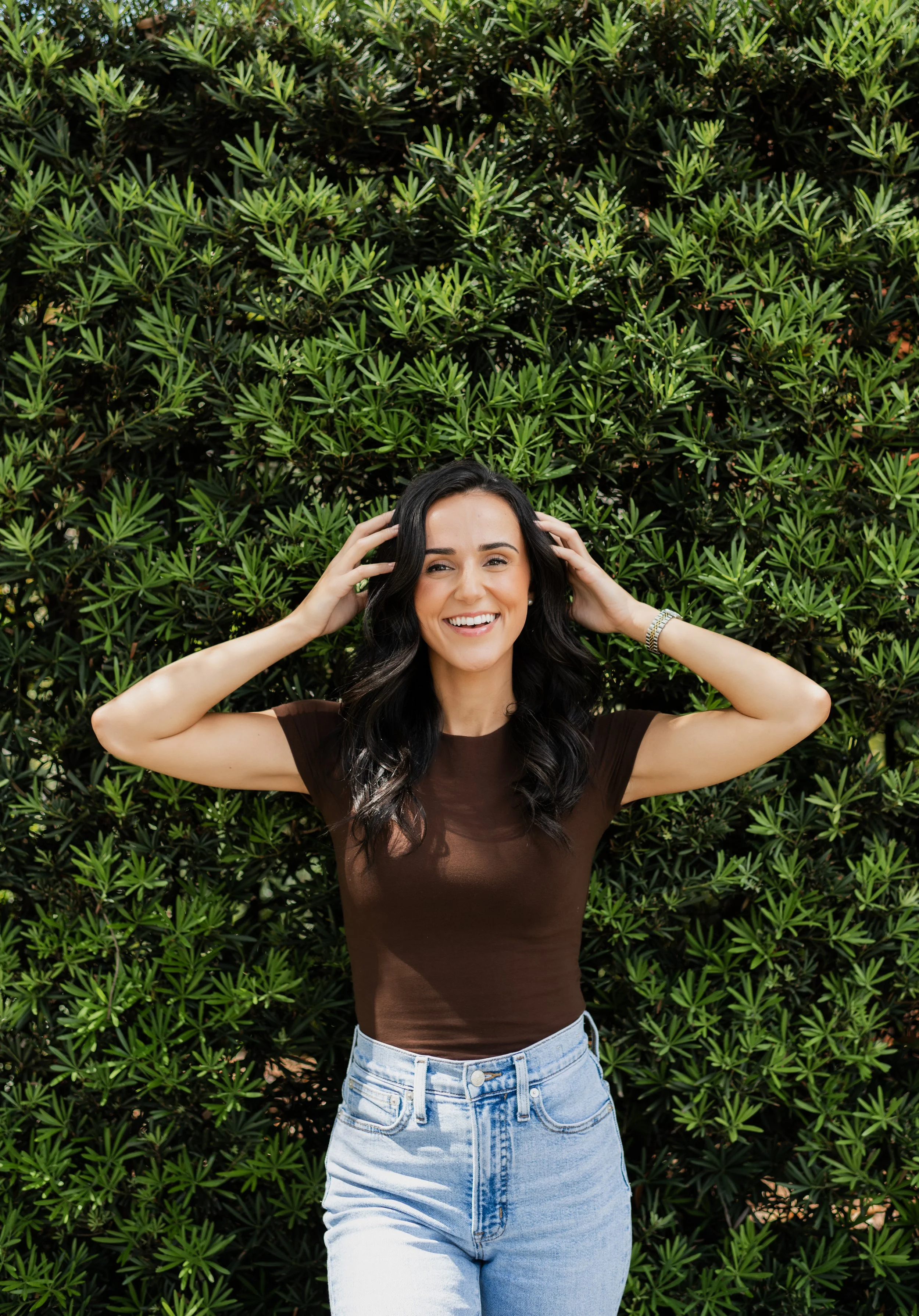 A woman with black wavy hair and a big smile, wearing a dark brown t-shirt and light blue jeans, standing in front of a lush, green leafy shrub, touching her hair with both hands.