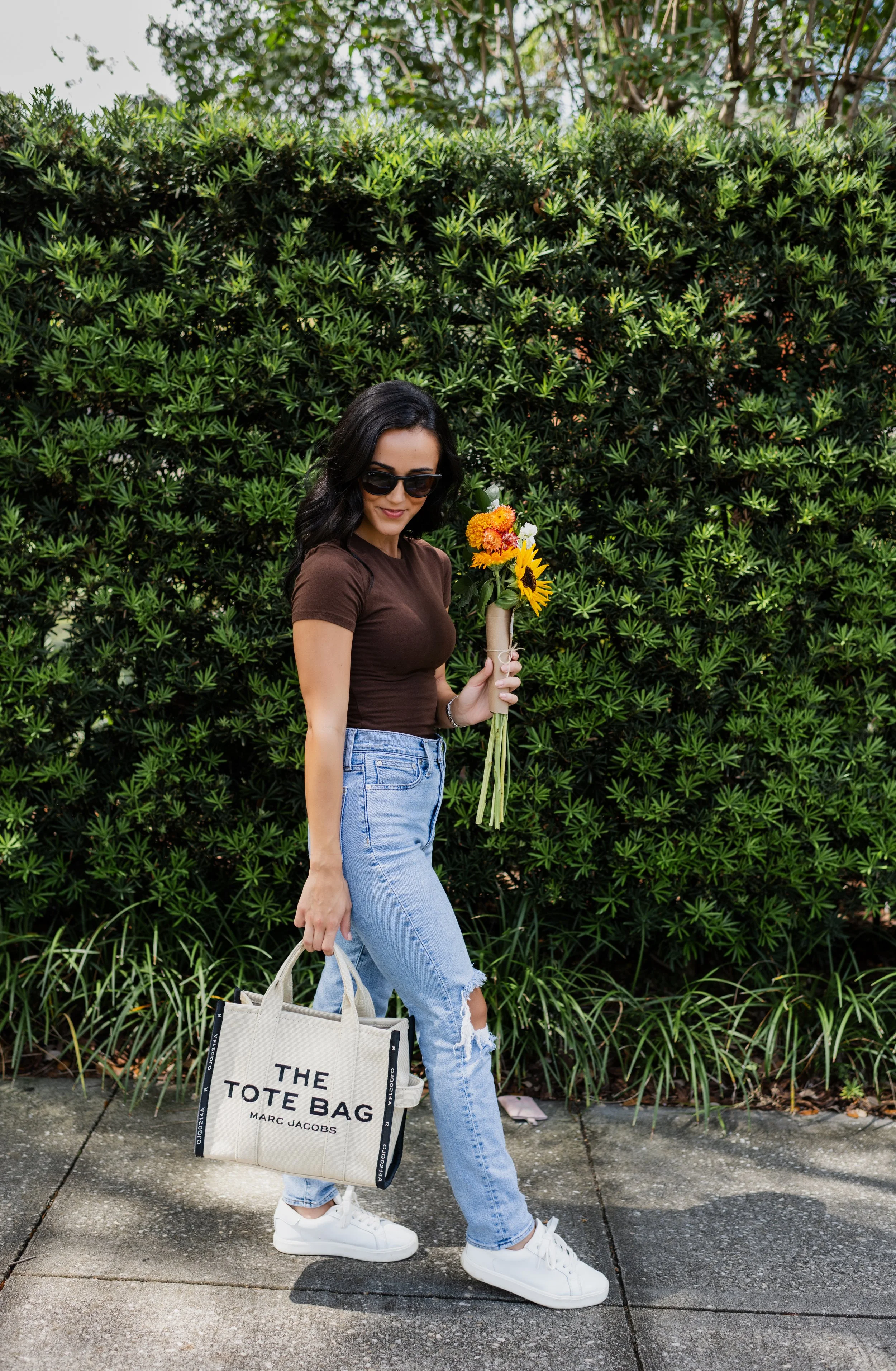 A woman with dark hair, sunglasses, wearing a brown t-shirt, light blue ripped jeans, and white sneakers, holding a bouquet of colorful flowers and a beige tote bag with black accents and the text "THE TOTE BAG" and "MARC JACOBS" printed on it, stand