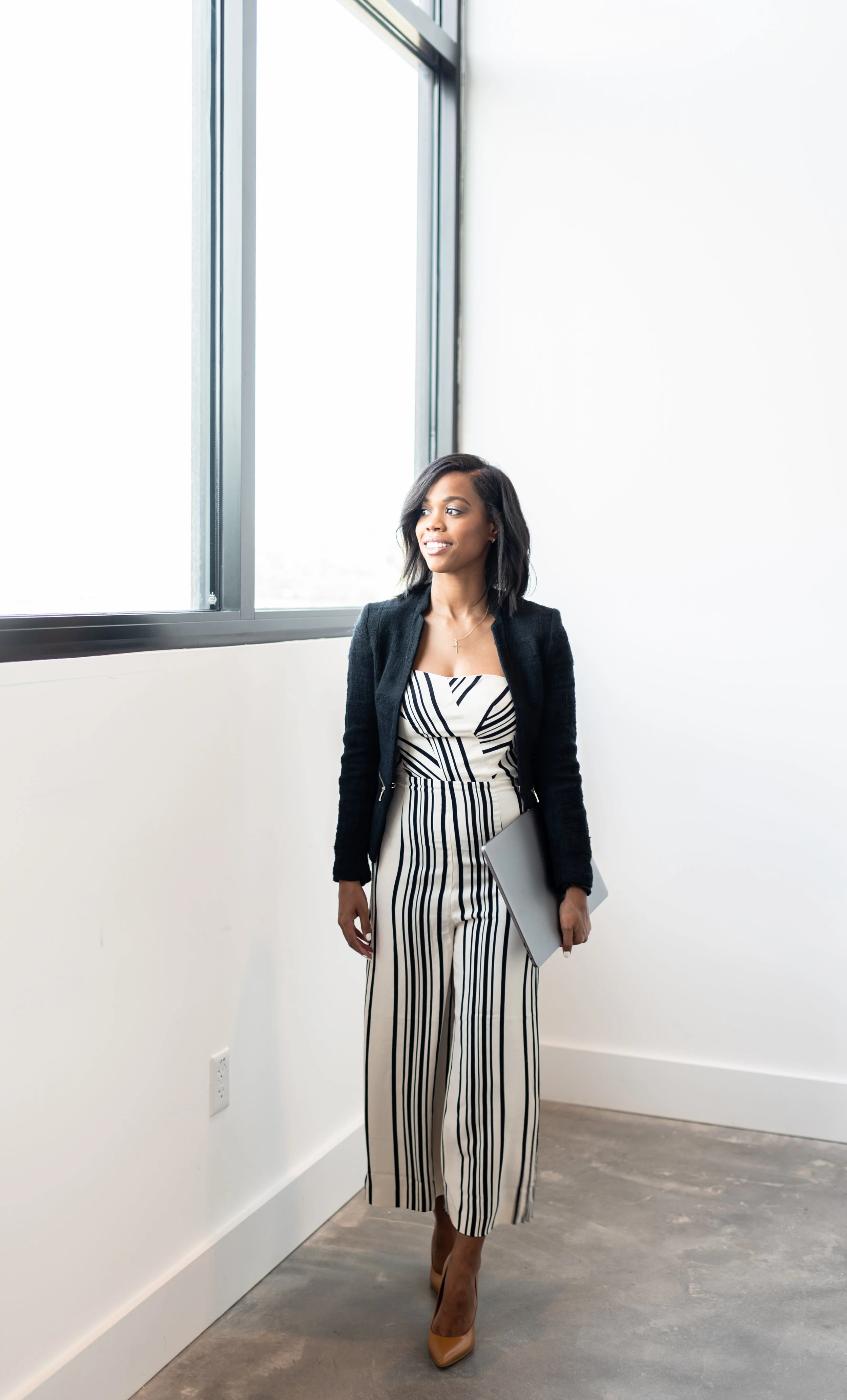 A woman in a black and white striped jumpsuit and black blazer walking in a bright, minimal office space with large windows, holding a gray laptop.