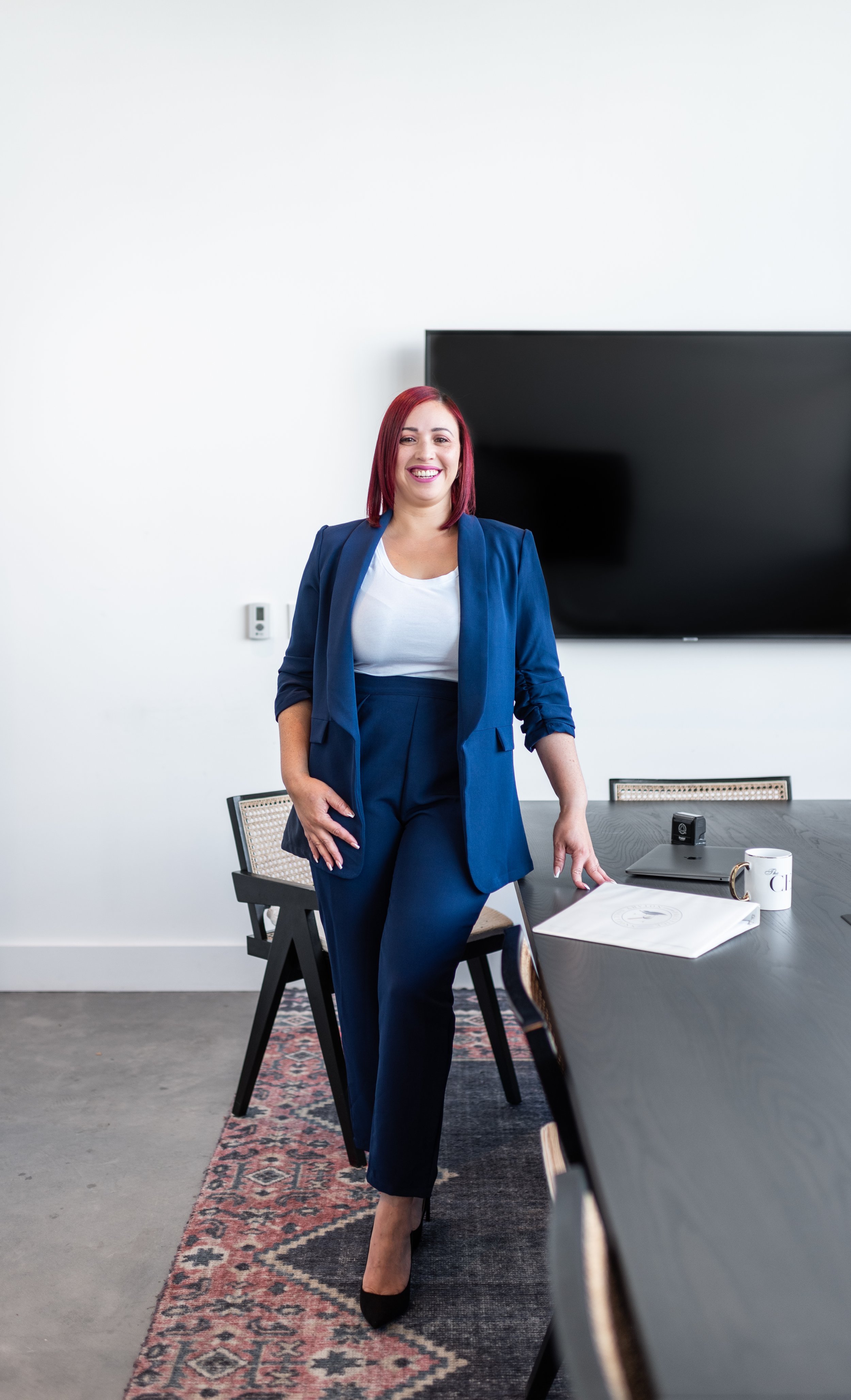 A woman with red hair in a blue suit standing in a conference room with a large TV, a laptop, a coffee mug, and a handout on the table.