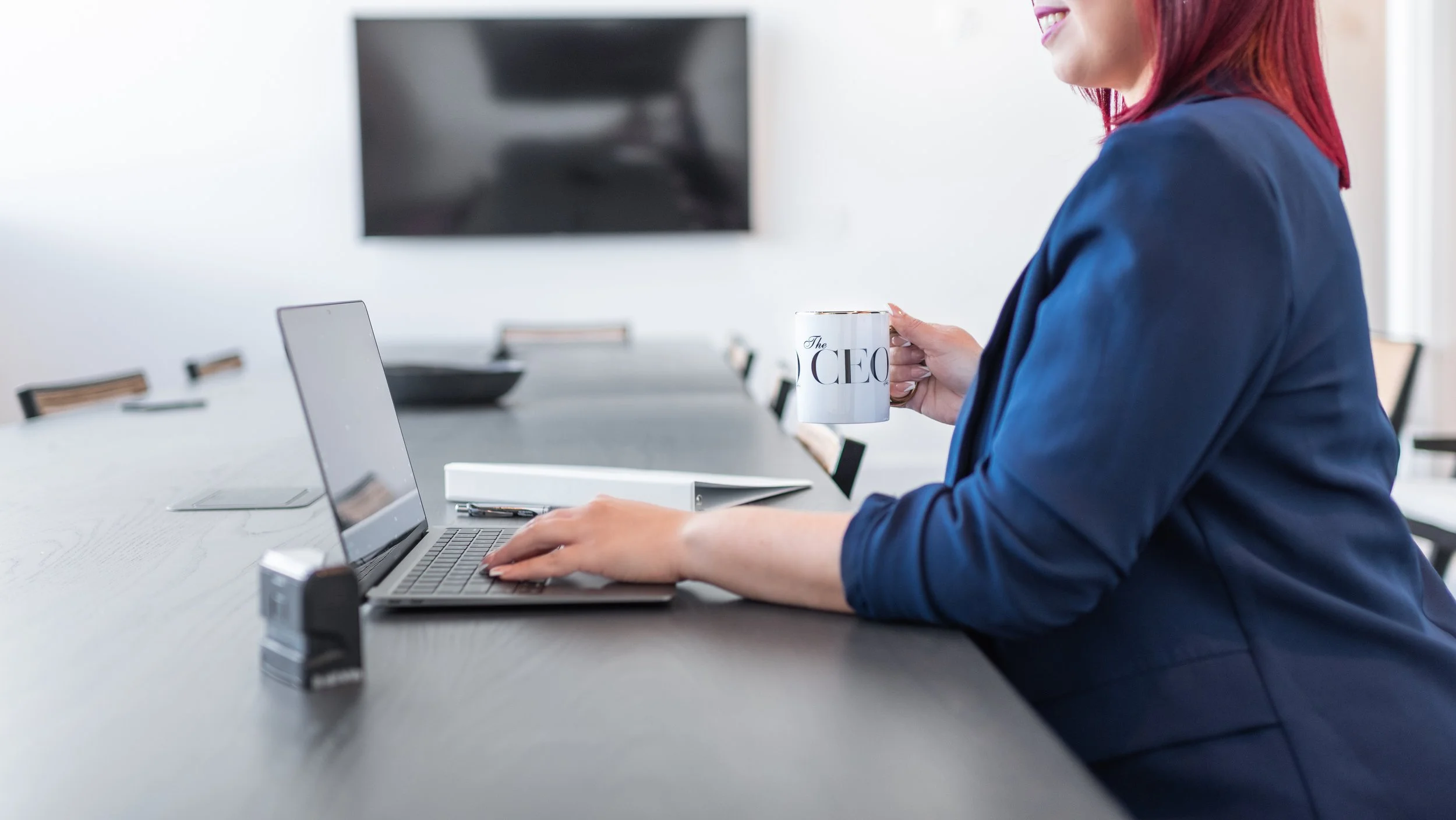 Businesswoman in blue blazer working on laptop at conference table, holding a white coffee mug with 'The CEO' text, in modern office setting.
