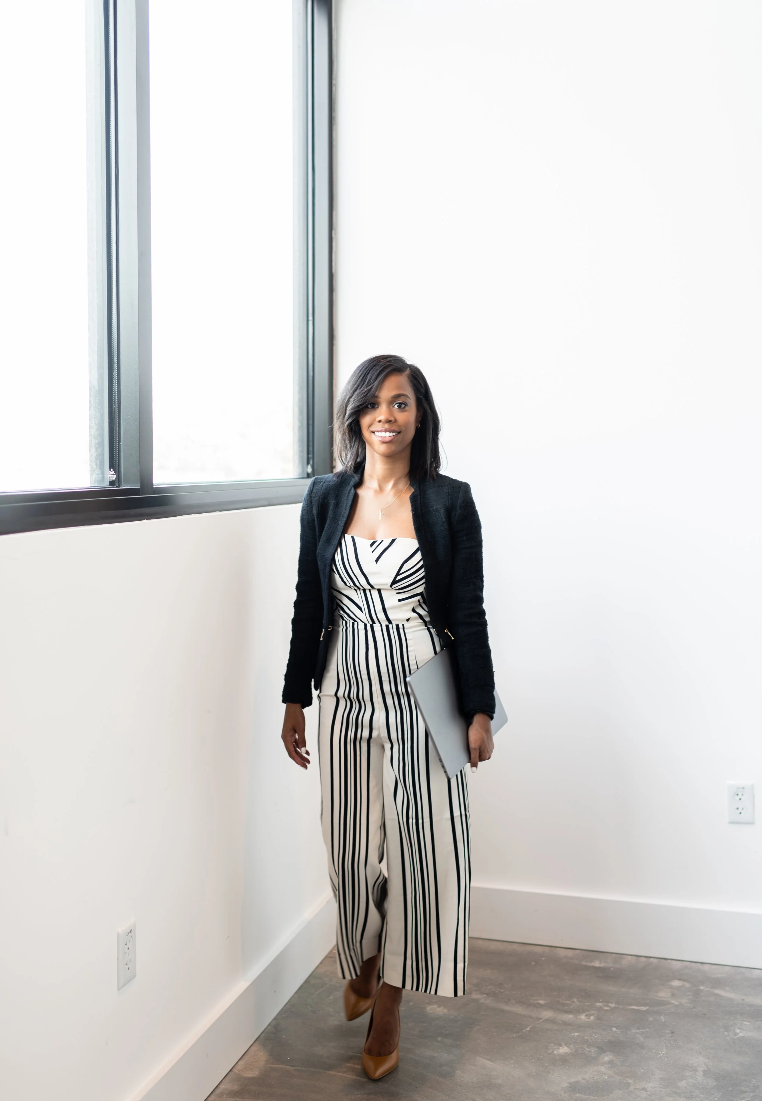 Woman standing indoors near a window, holding a laptop, dressed in a striped jumpsuit with a black blazer and tan heels.