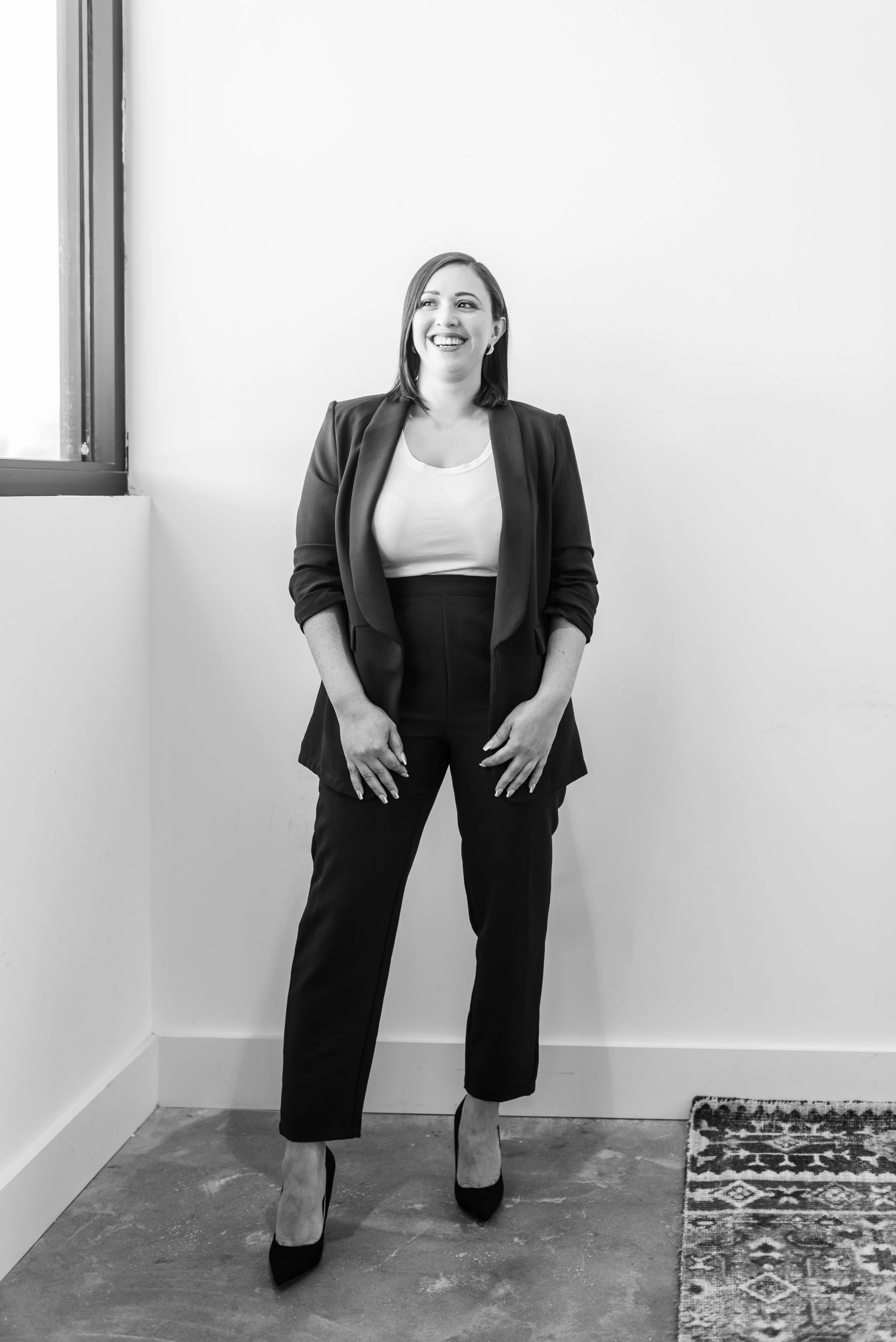 Black and white photo of a woman in a blazer and pants, smiling and standing near a corner and window