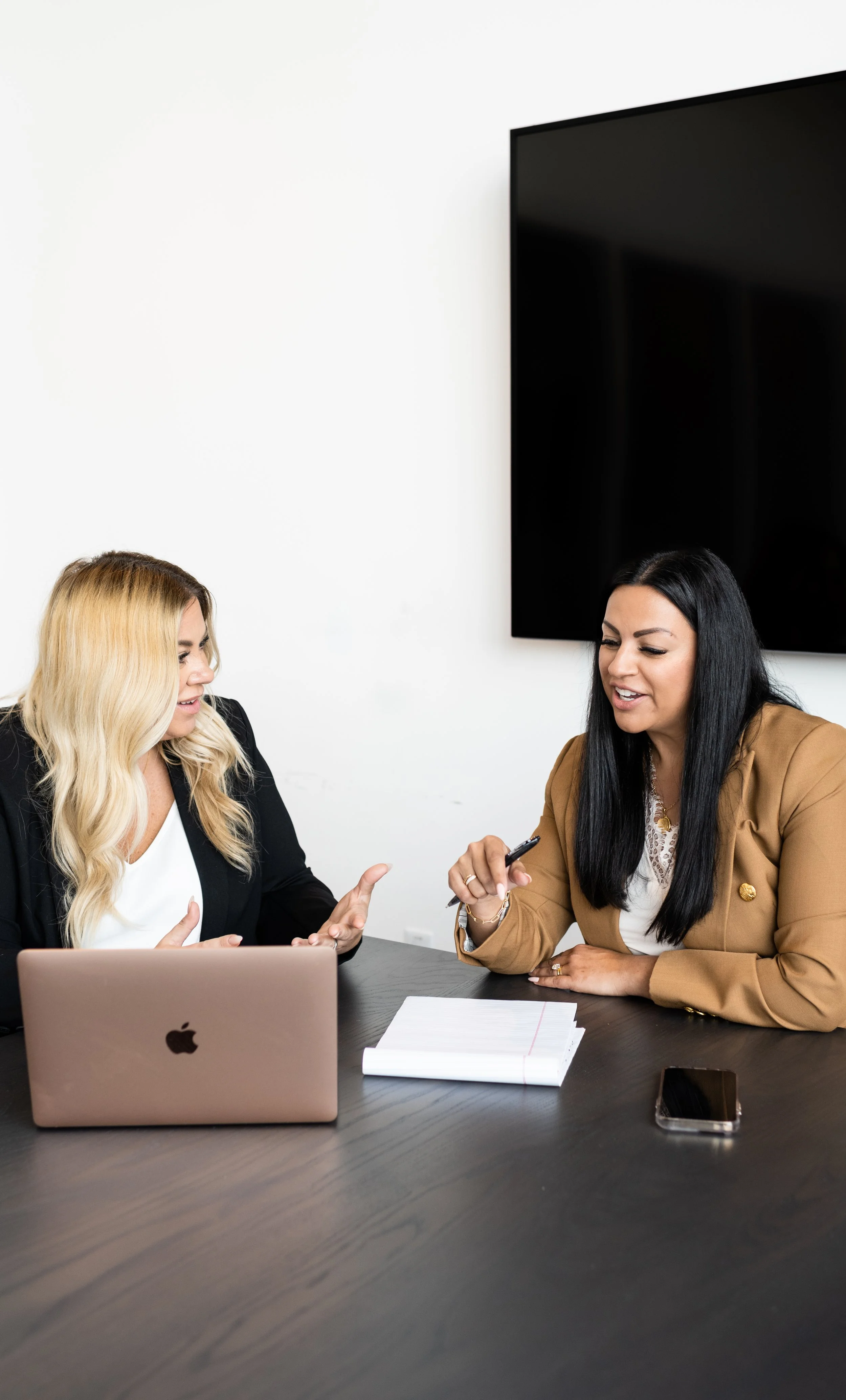 Two women are sitting at a desk in a meeting room, engaged in conversation. One woman has blonde hair and is wearing a black blazer, the other has black hair and is wearing a tan blazer. There is a silver laptop, a notebook, and a smartphone on the d