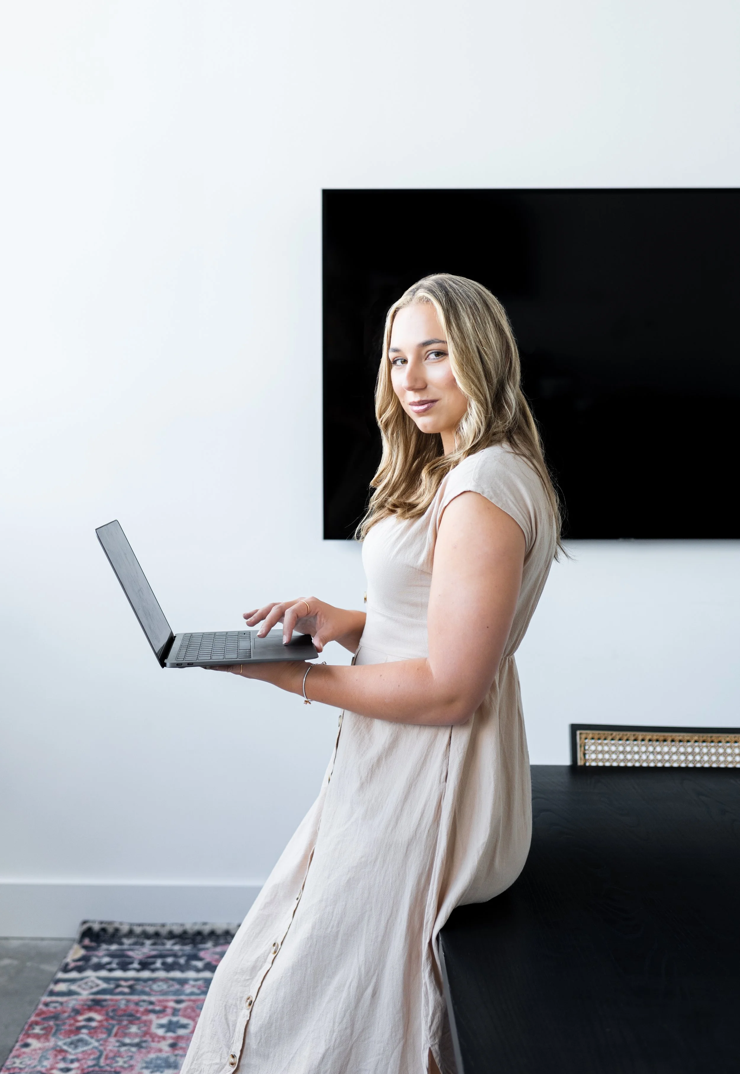 A woman with blonde hair in a beige dress sitting on a black table, using a laptop, in a room with white walls and a flat-screen TV behind her.