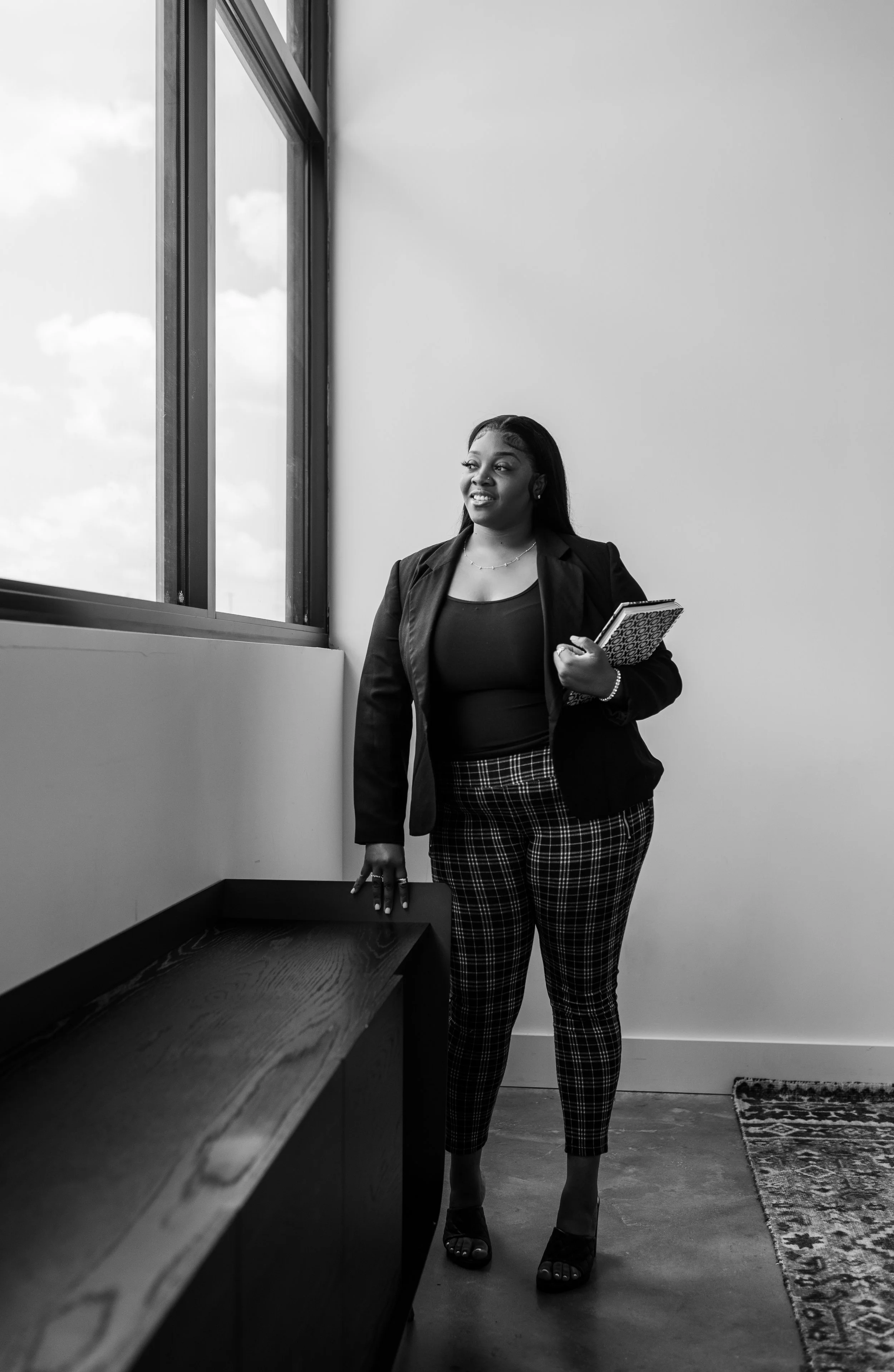 A woman standing in a modern office or meeting room near a large window, holding notebooks and dressed in business attire.