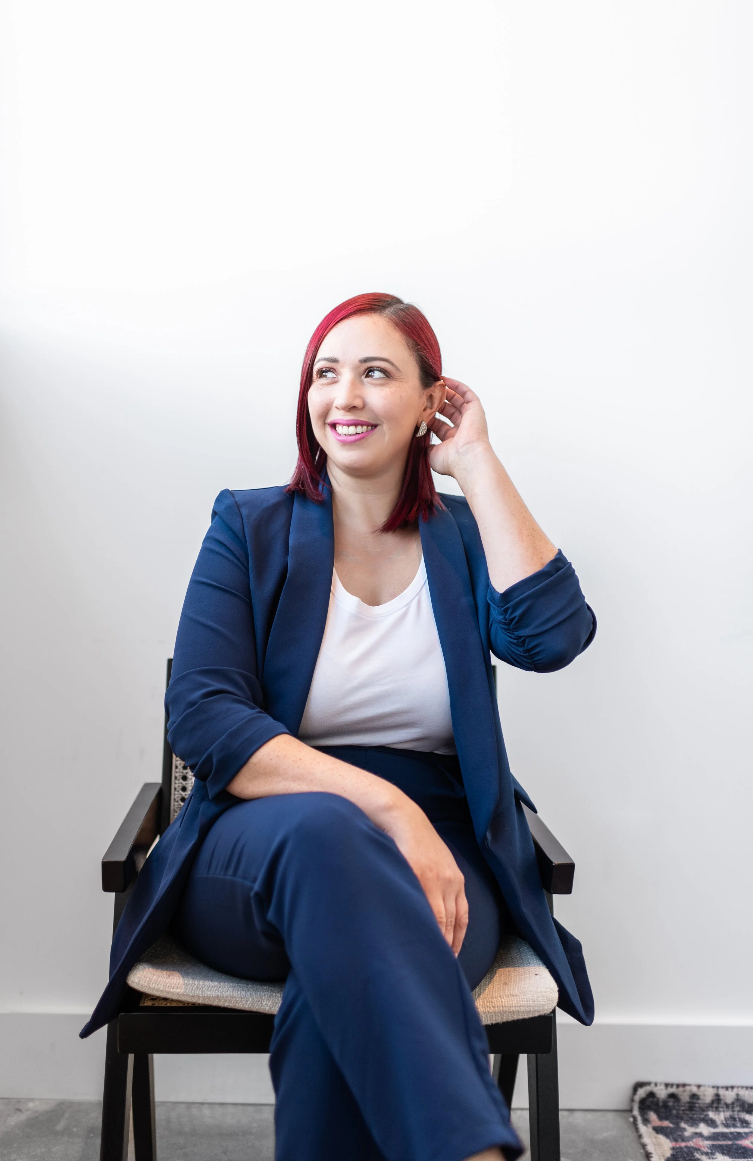 A woman with red hair, dressed in a blue blazer and pants, sitting on a chair with a white background, smiling and touching her ear.
