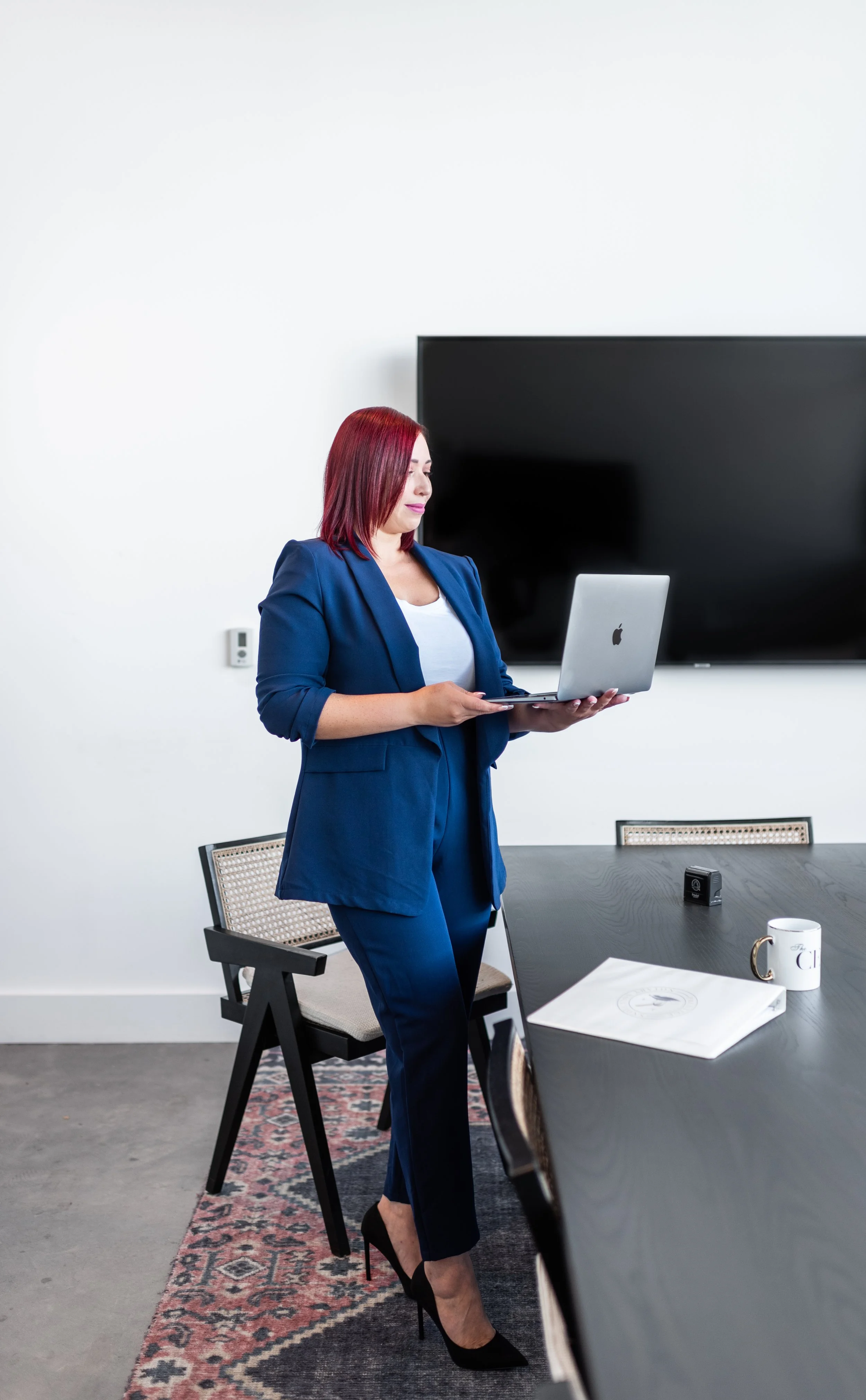 A woman in a blue pantsuit and heels standing in an office, looking at a laptop with a large TV screen behind her.