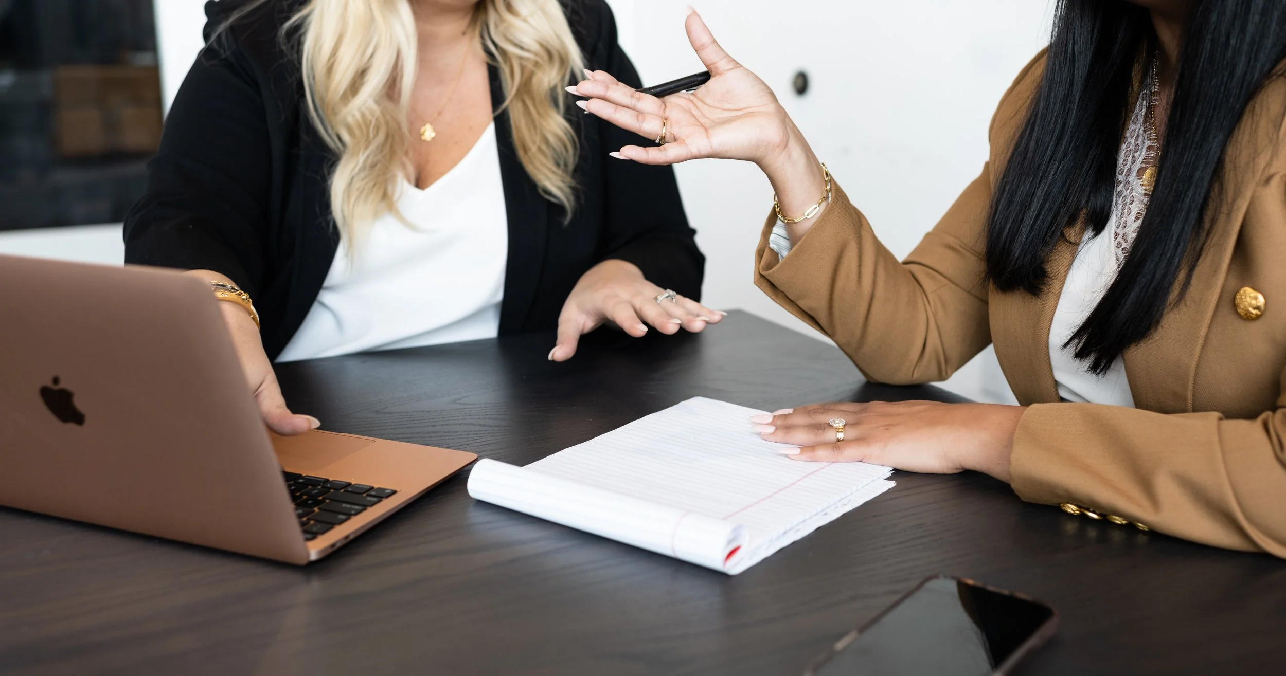 Two women having a discussion at a desk with a rose gold MacBook, notebook, and phone.