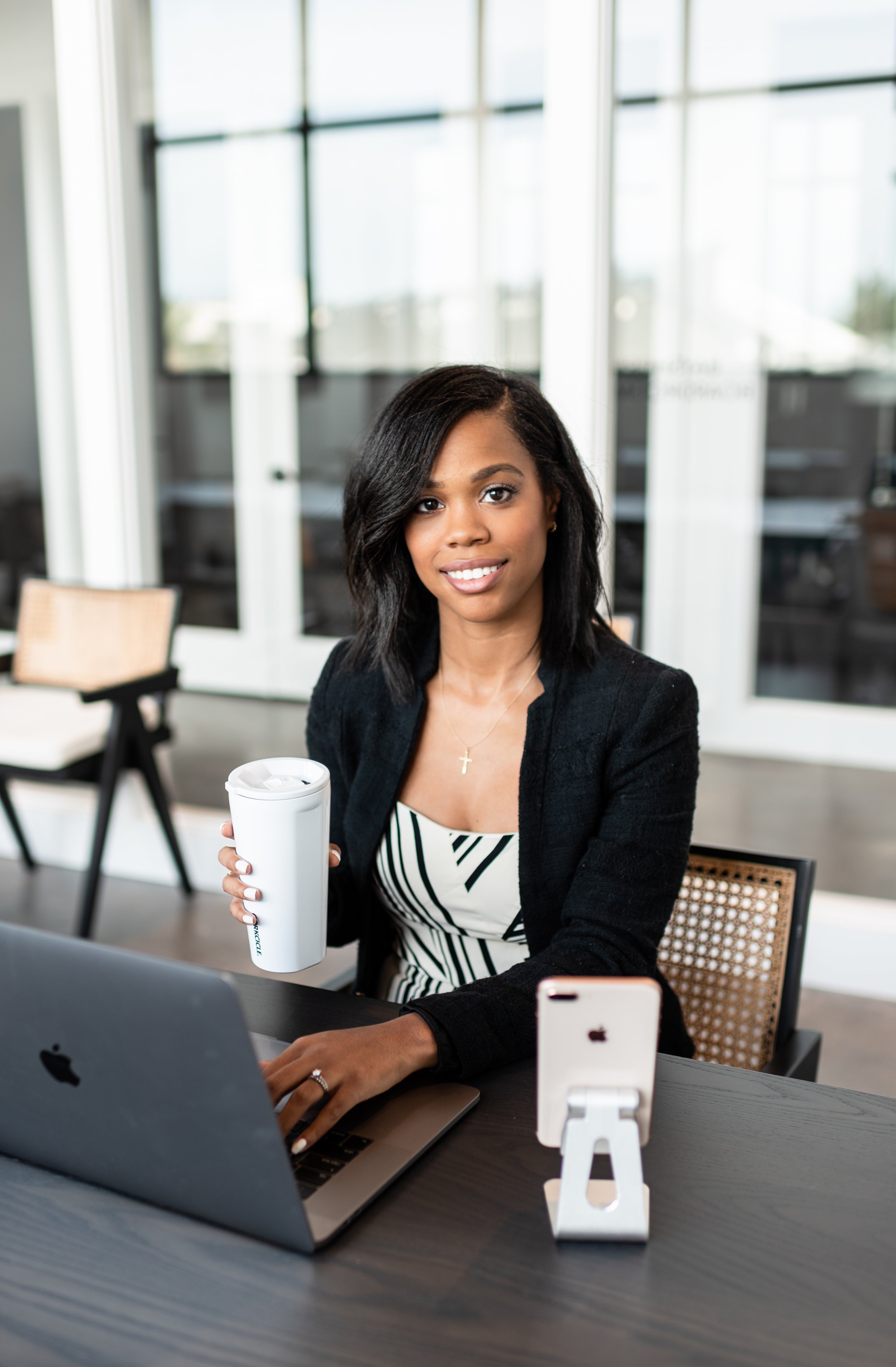 A woman sitting at a desk with a laptop, holding a white tumbler, in a modern office space with glass windows.