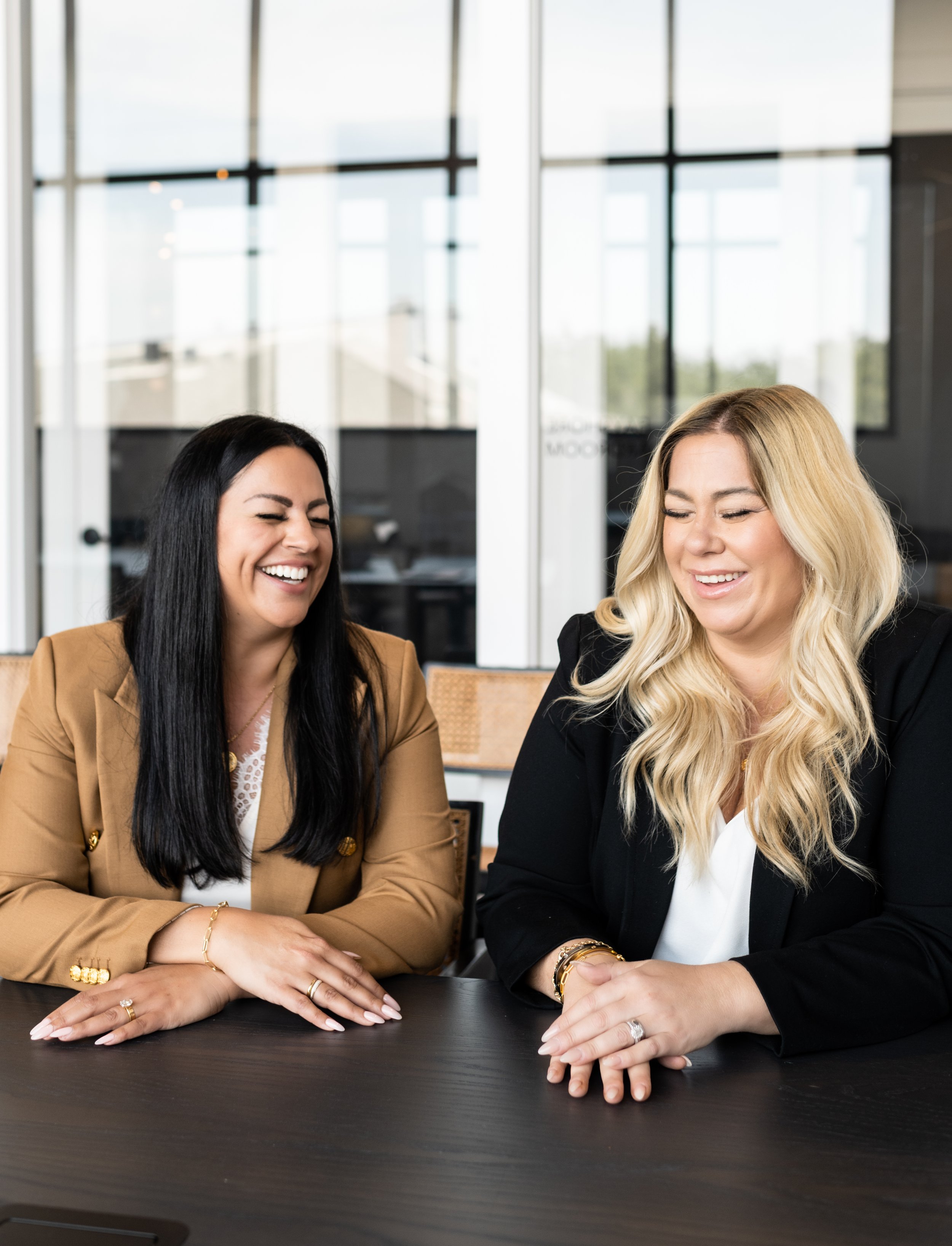 Two women with light skin smiling and laughing at a table in an office setting.