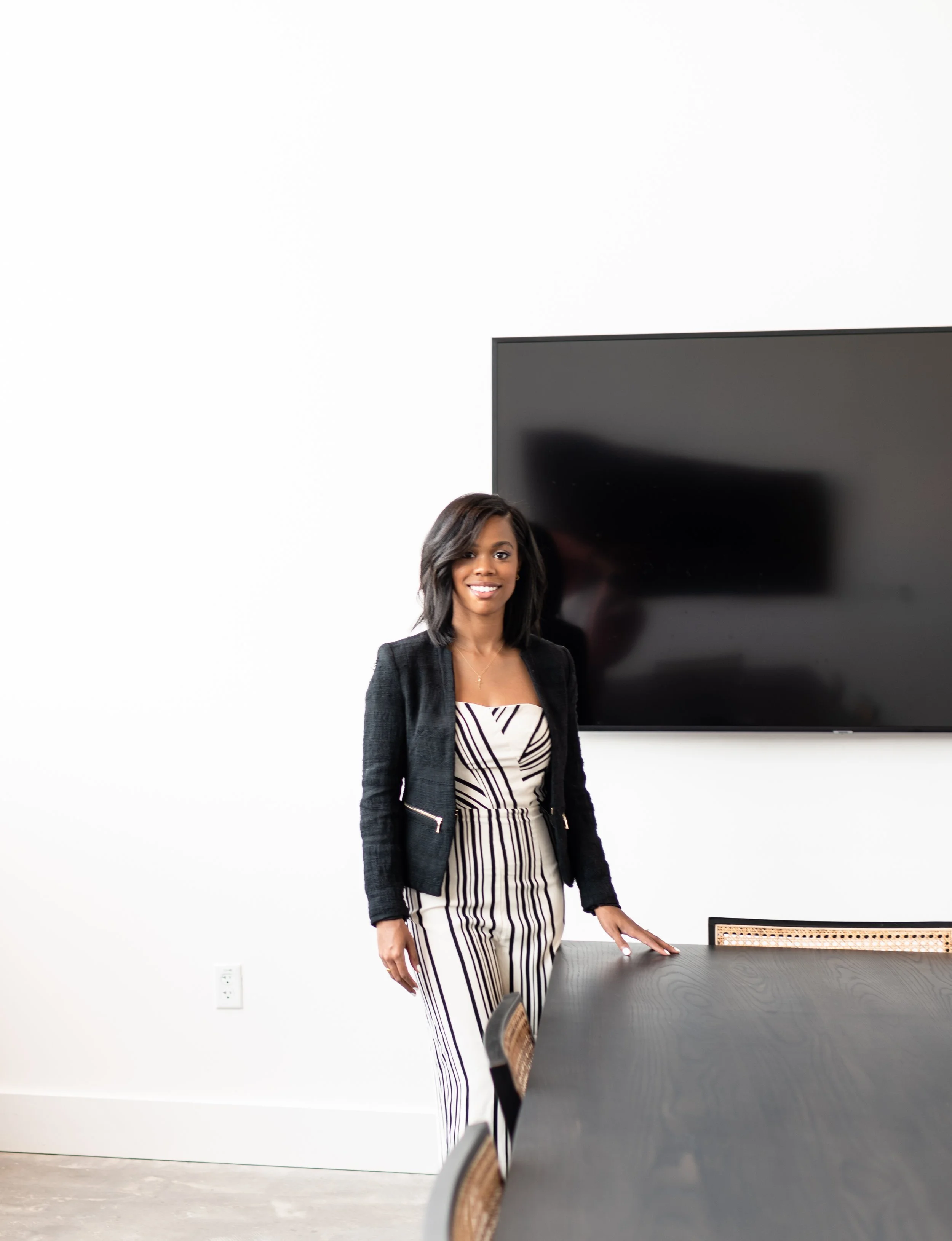 A woman with shoulder-length hair, wearing a black blazer and a striped jumpsuit, standing in a conference room and smiling at the camera.