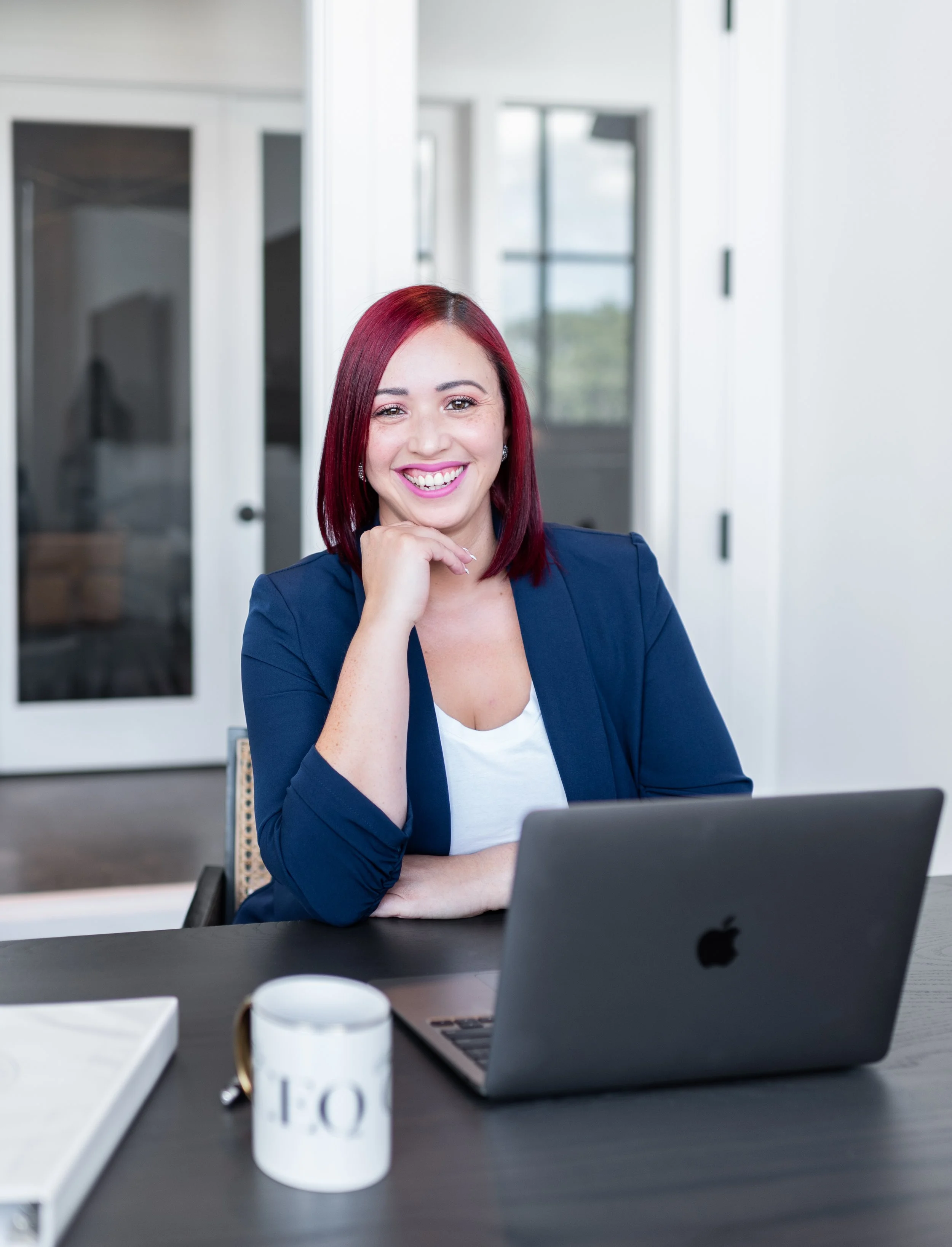 A woman with red hair, wearing a navy blazer and white top, sitting at a desk with an open laptop and mug, smiling at the camera in a modern office.