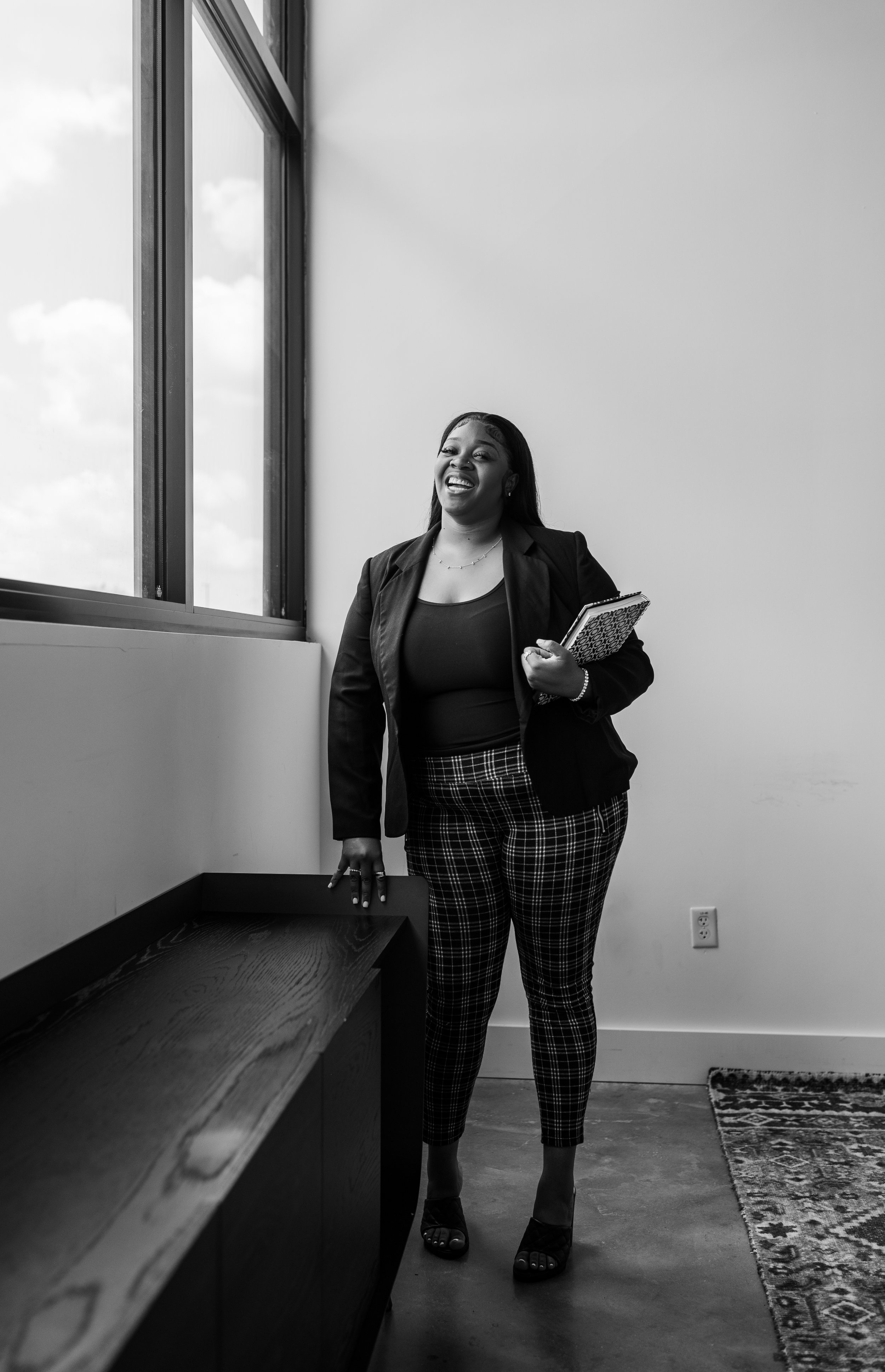 A woman stands in an office next to a window, smiling and holding a notebook, wearing a blazer, patterned pants, and open-toed shoes.