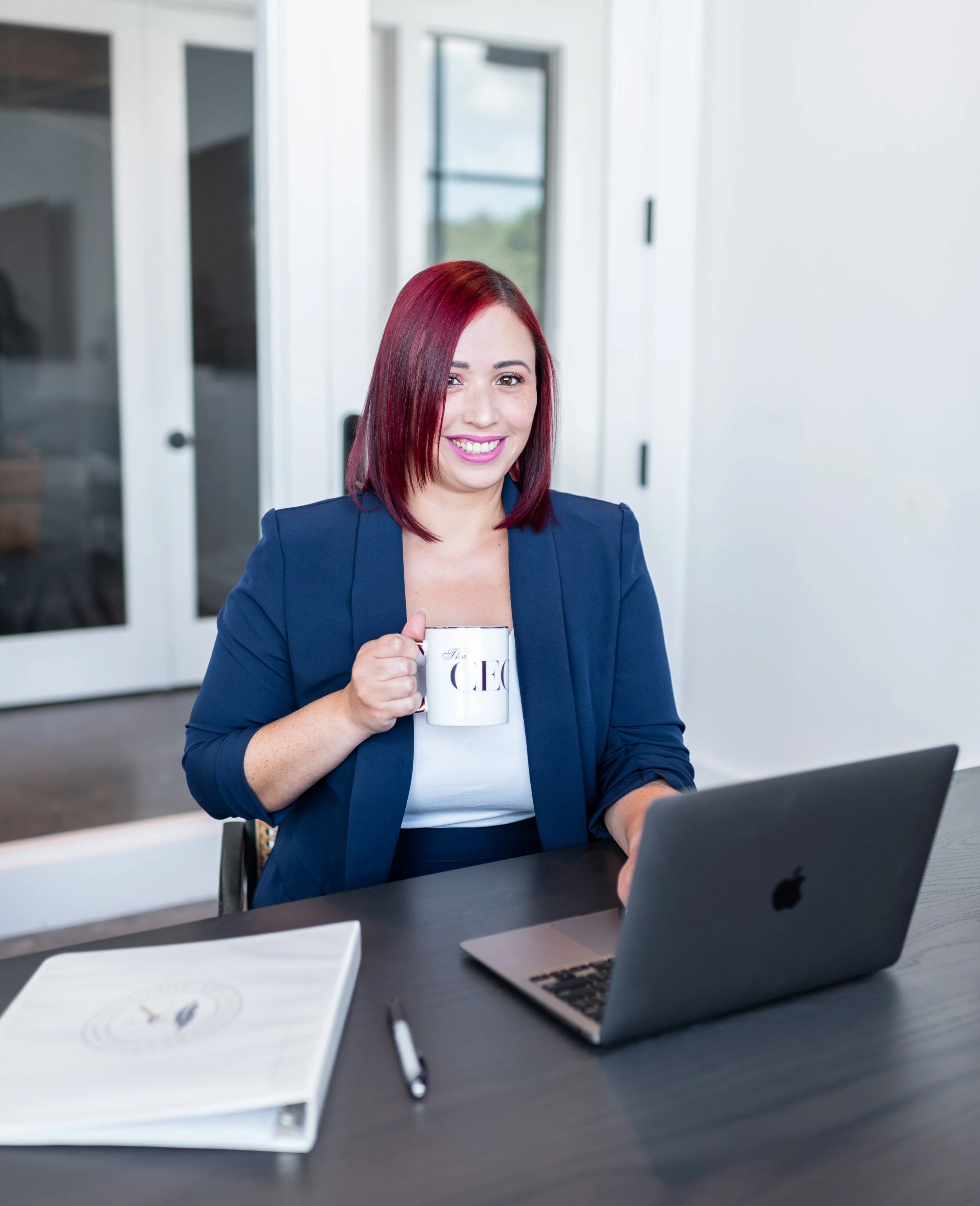 A woman with red hair, smiling, sitting at a desk with a MacBook, holding a mug that says 'The CEO', with a notebook and pen on the desk, in a modern office with glass doors.