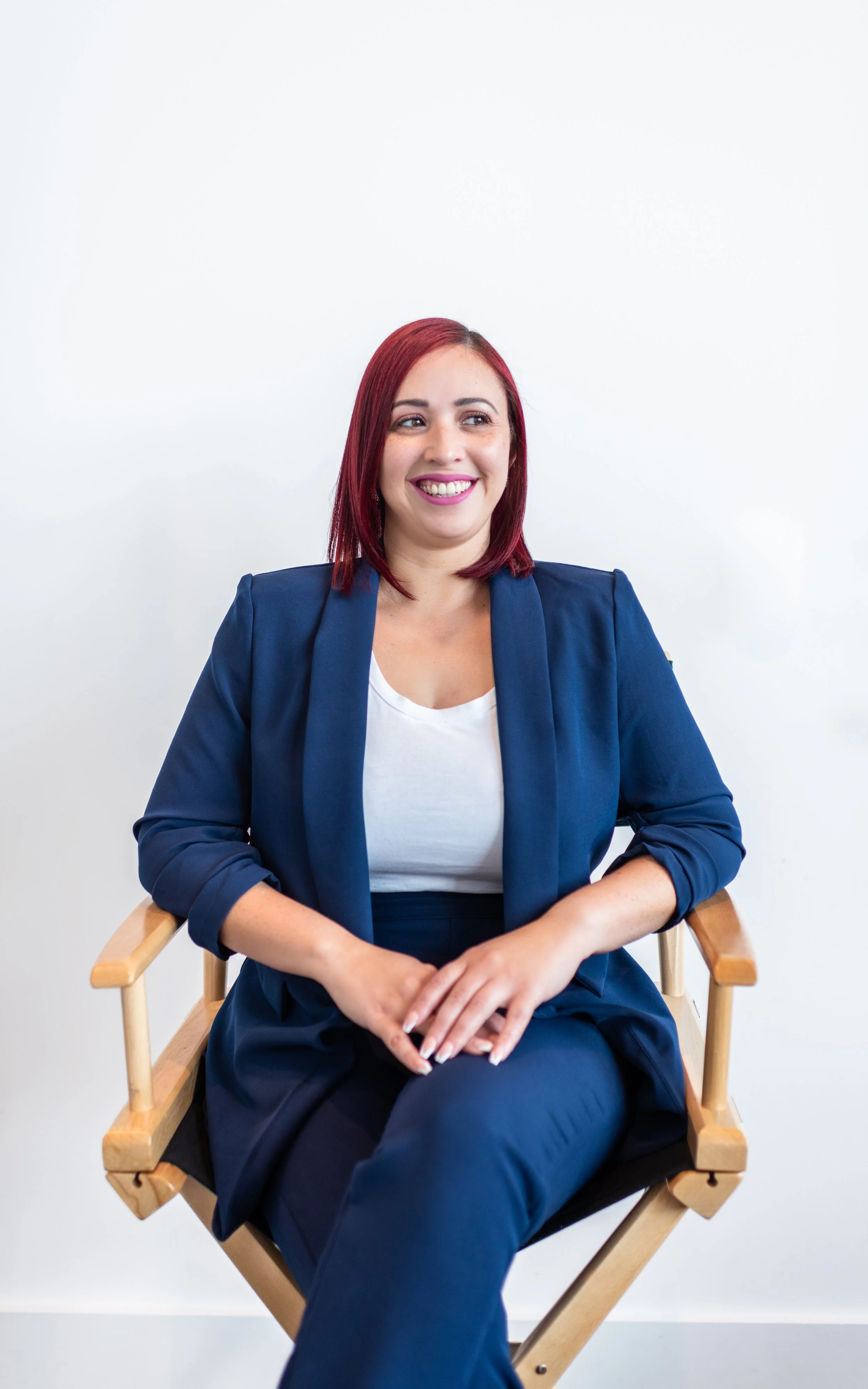 A woman with shoulder-length red hair, wearing a navy blazer and white top, sitting on a wooden director's chair, smiling and looking to her left, against a plain white background.
