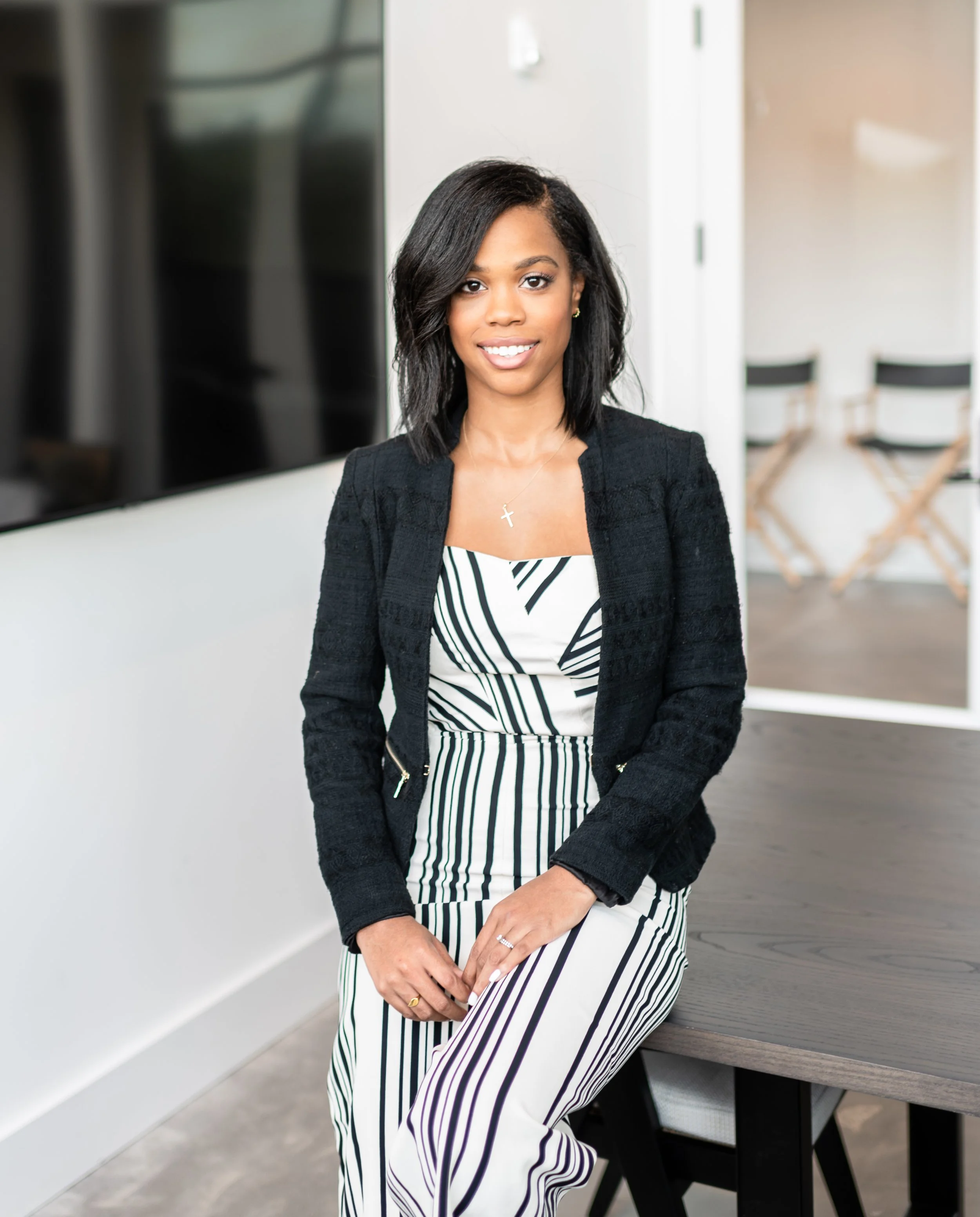 A woman in a black blazer and striped dress sitting in a modern office space.