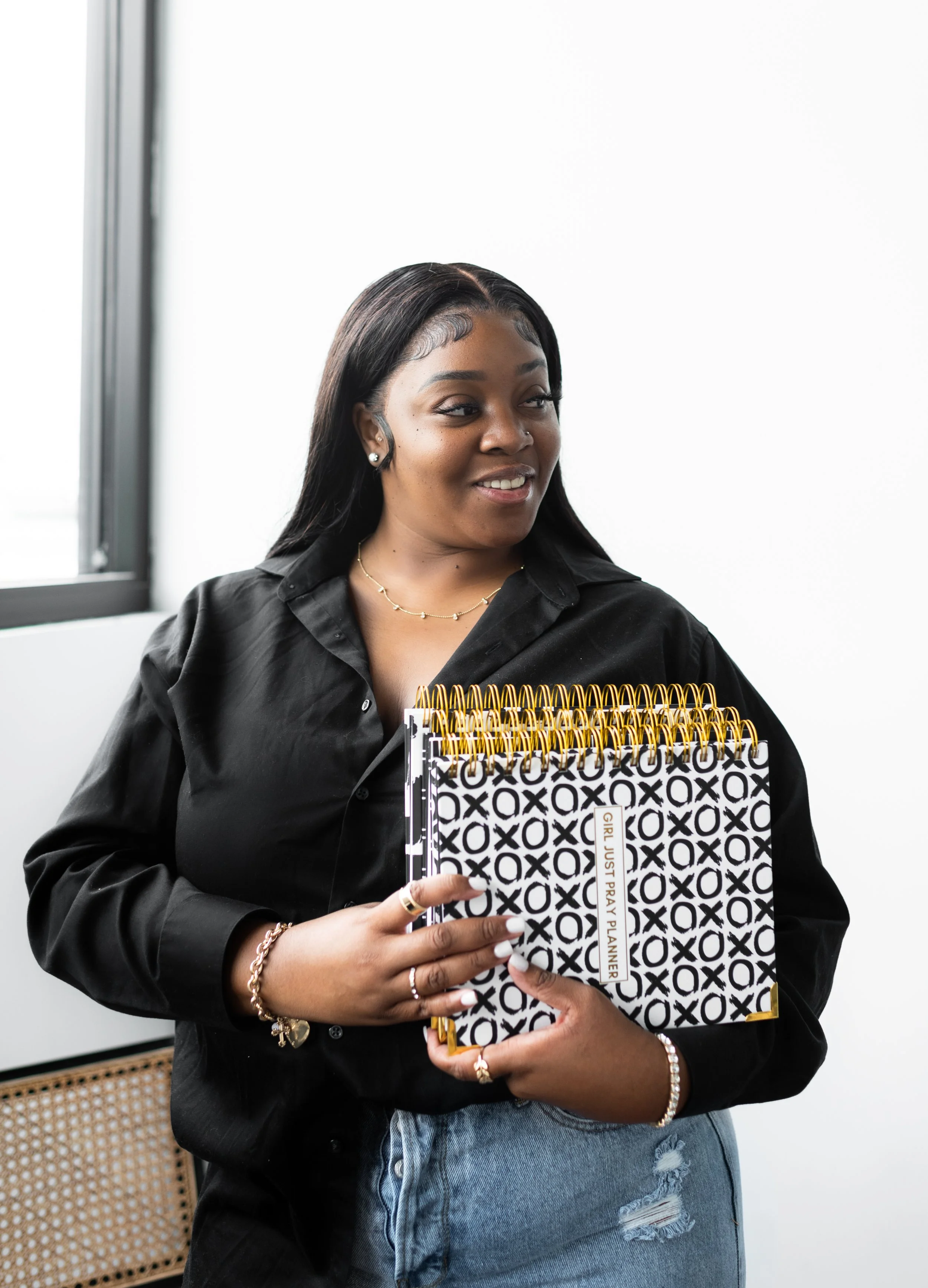 A young woman with long black hair, wearing a black shirt and jeans, holding several black and white patterned planners with gold coils, standing in a bright room with a white wall and a window in the background.
