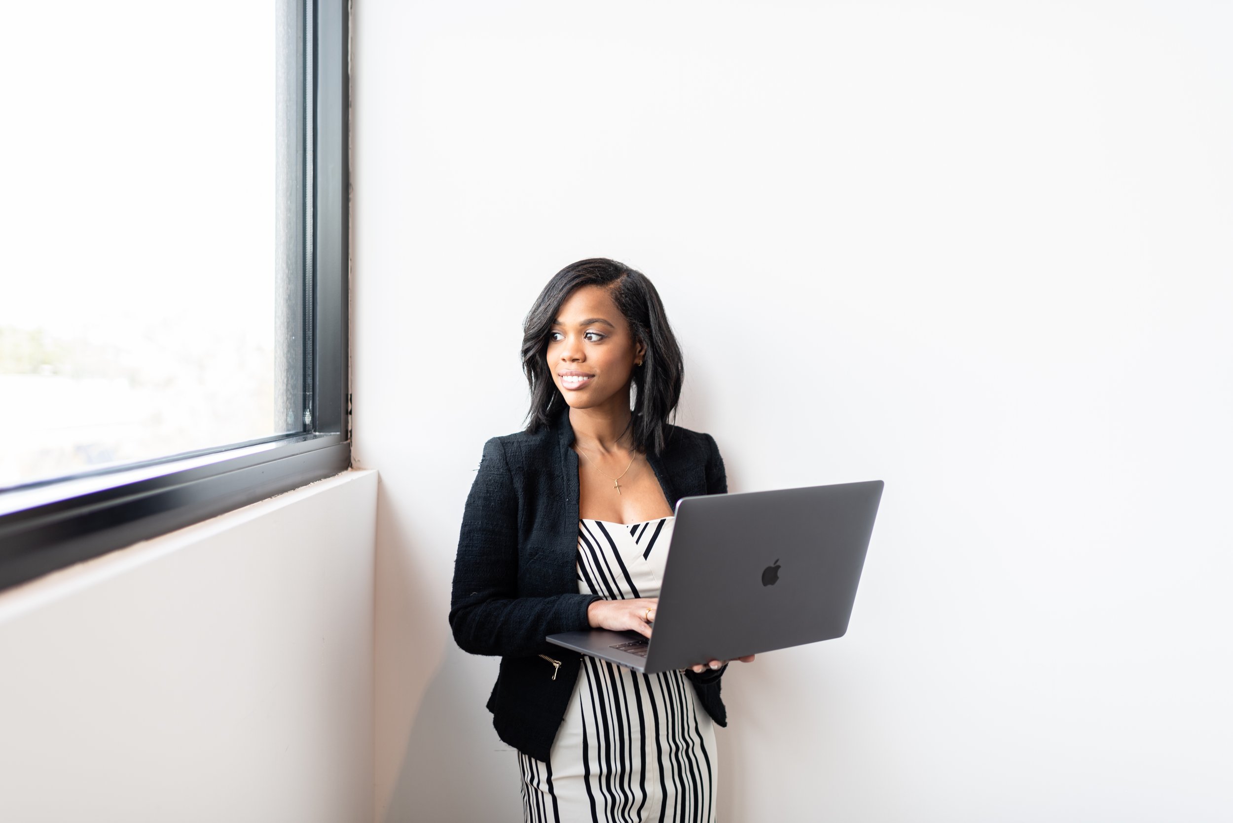 A young woman with dark hair wearing a black blazer and a black-and-white striped dress holding a MacBook laptop and standing next to a window.
