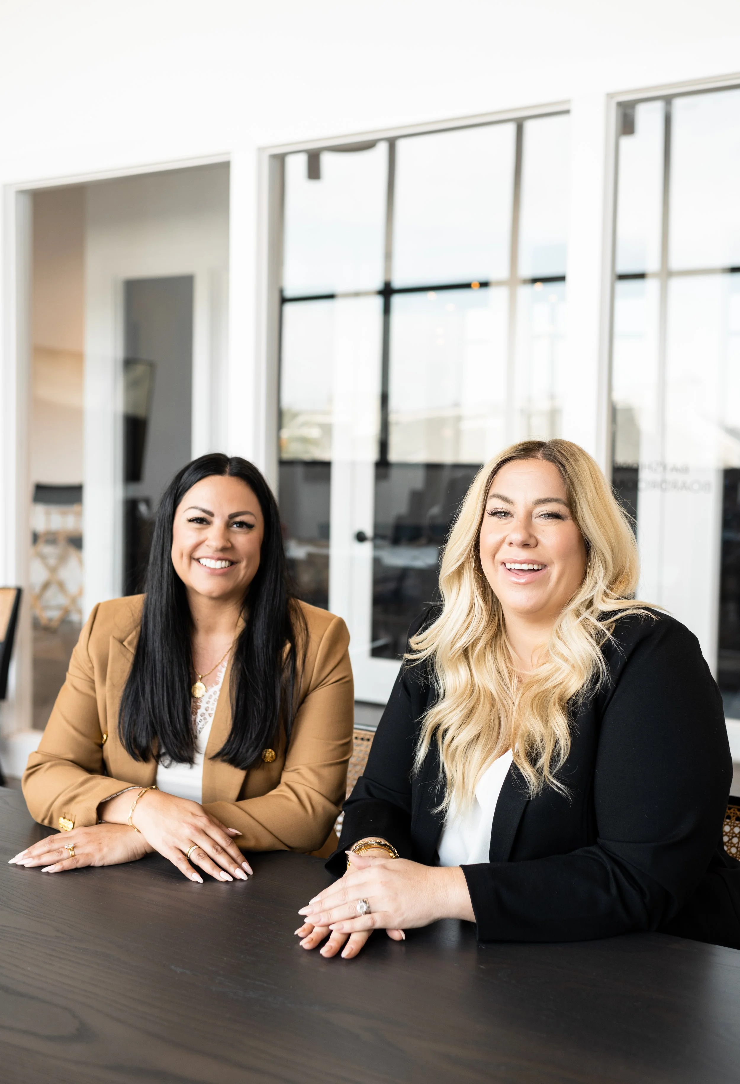 Two women sitting at a table in a modern office, smiling at the camera.