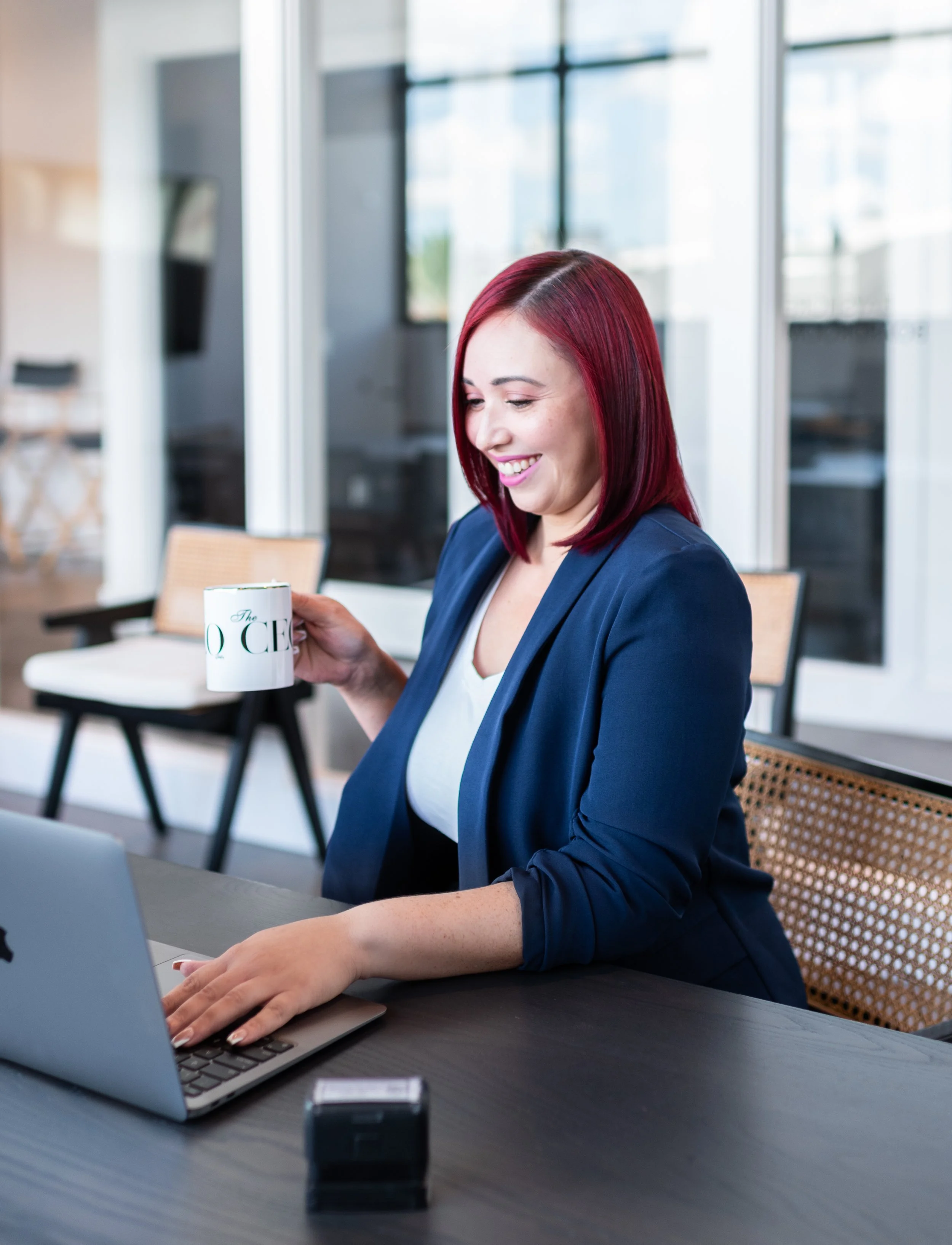 A woman with red hair in a business suit smiling and holding a coffee mug while working on a laptop at a desk in a bright modern office.