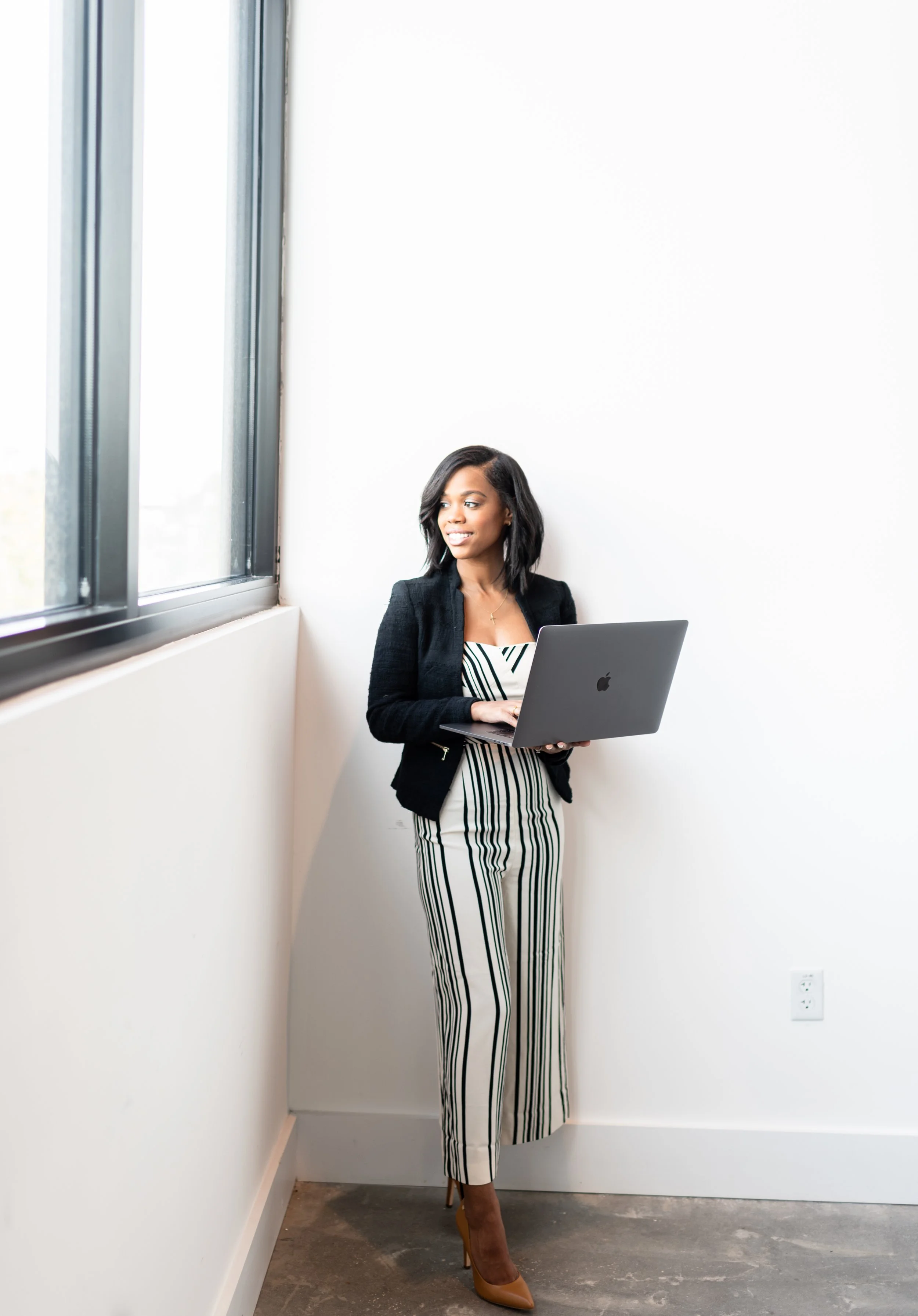 A woman with black hair in a bob hairstyle holding an open silver laptop while standing by a large window in a white-walled room, wearing a black blazer, striped pants, and tan high heels.