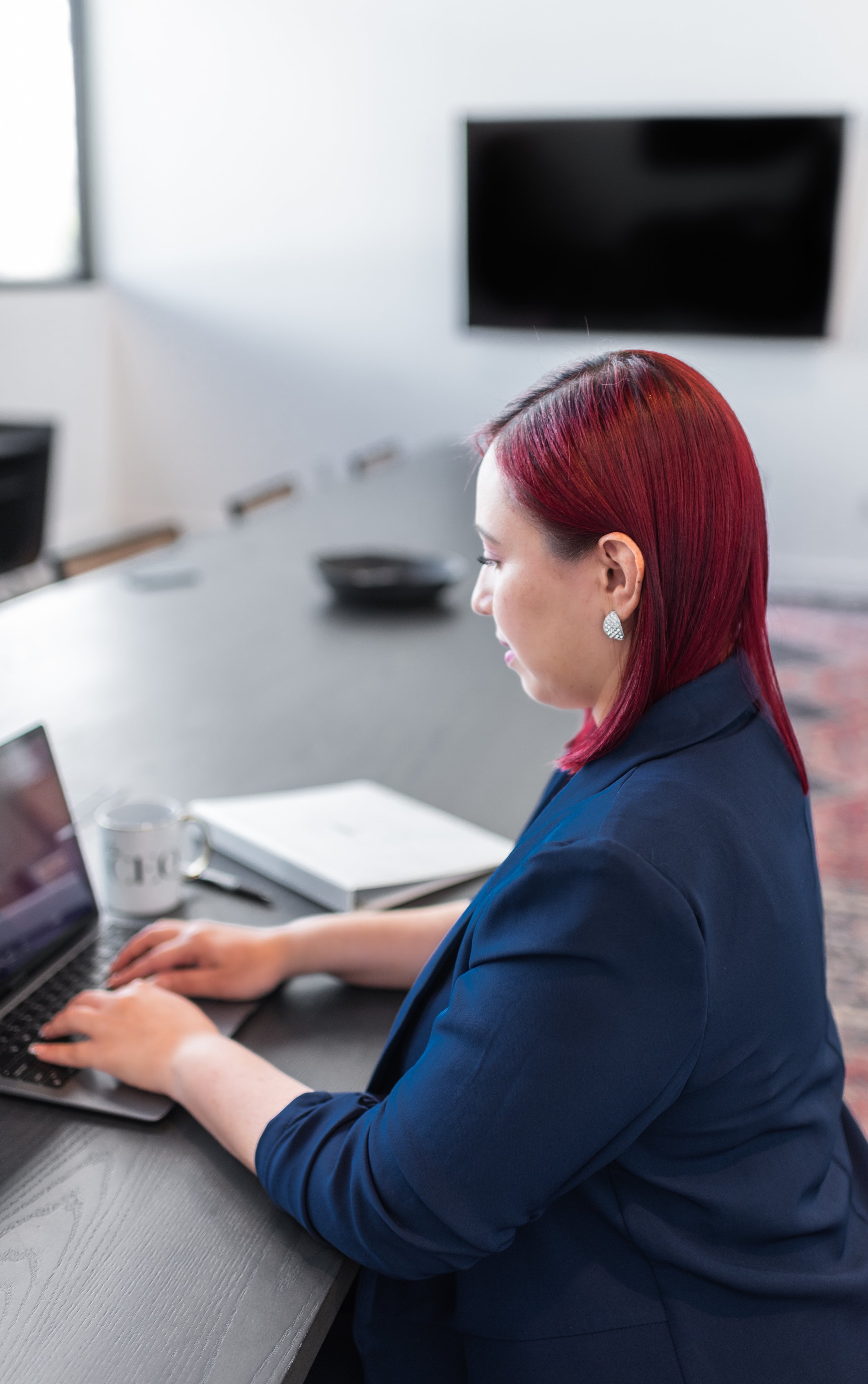A woman with red hair working on a laptop in a conference room, with a coffee mug, notebook, and pen on the table, and a black bowl in the background.
