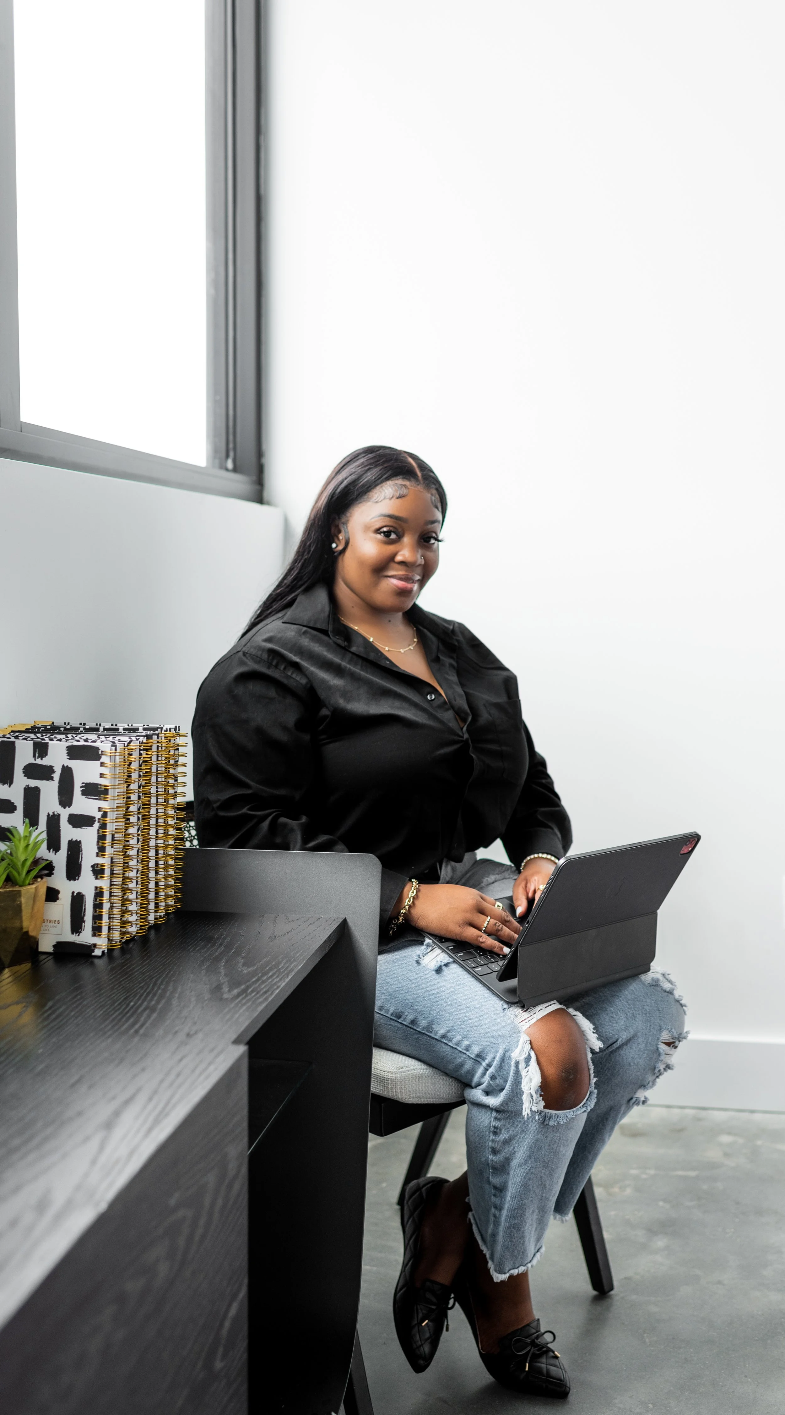 A woman sitting at a desk with a laptop, wearing a black shirt and ripped jeans, smiling at the camera in a modern office setting.