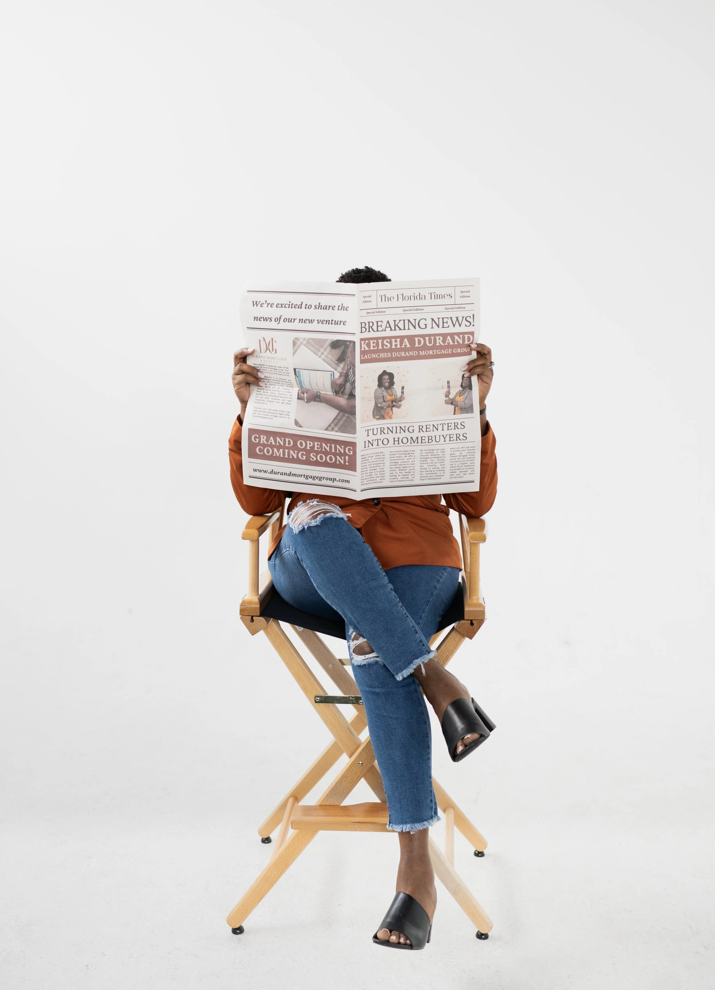 A woman sitting on a wooden director's chair, reading a newspaper against a plain white background, with her legs crossed and wearing a rust-colored top, ripped blue jeans, and black high-heeled sandals.