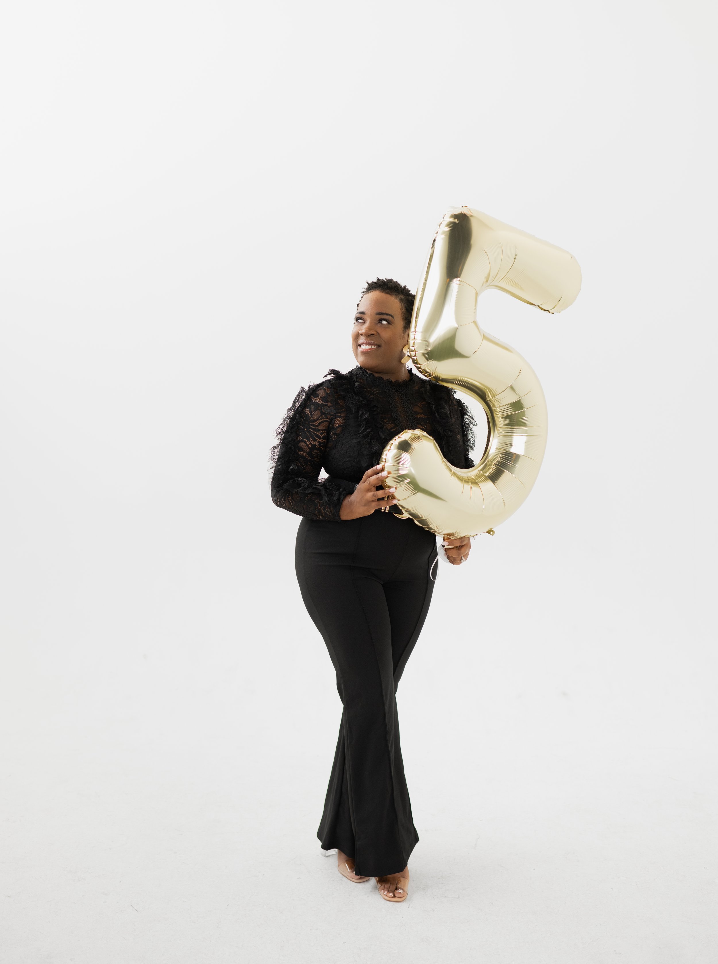 A woman in black lace top and black pants holding a large gold balloon in the shape of the number five, standing against a plain white background.