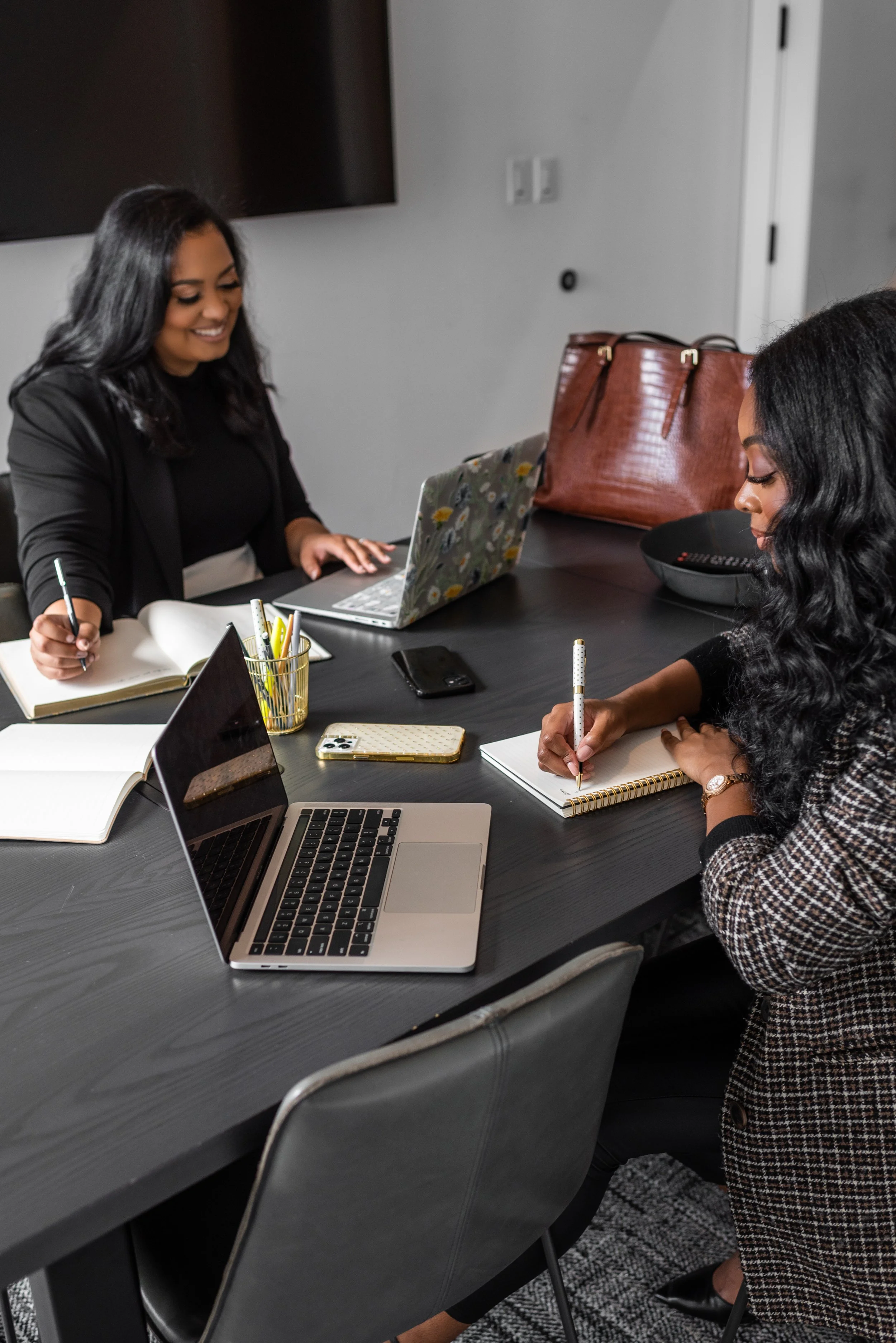 Two women working together at a conference table with laptops, notebooks, and pens during a business meeting.