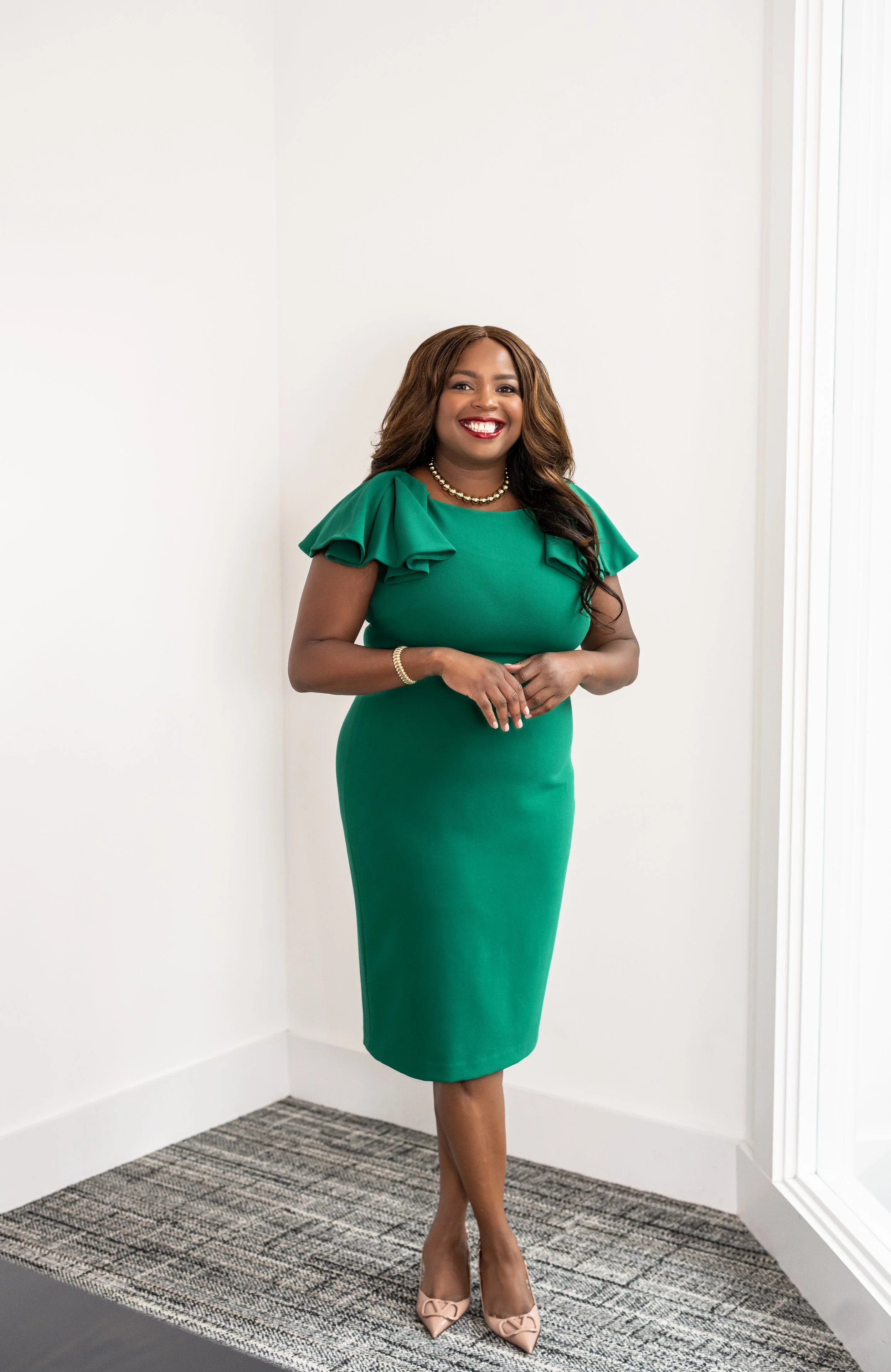 A woman with brown hair in loose curls wearing a green dress with puff sleeves, beige high heels, gold jewelry, and smiling, standing in a white room with natural lighting.