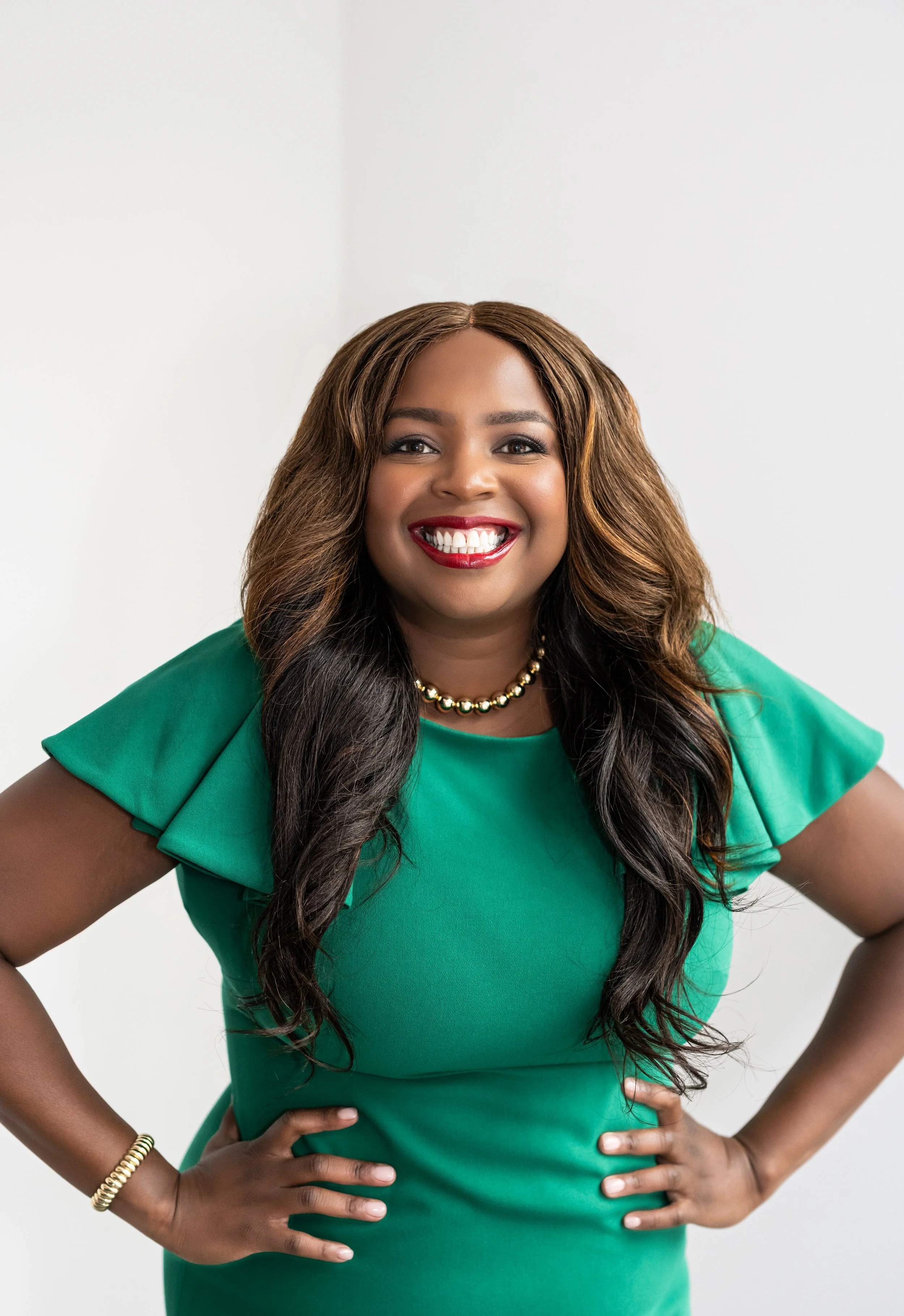A woman with long, wavy dark hair, wearing a green dress with ruffled sleeves, a pearl necklace, and a gold bracelet, smiling with hands on her hips against a plain white background.