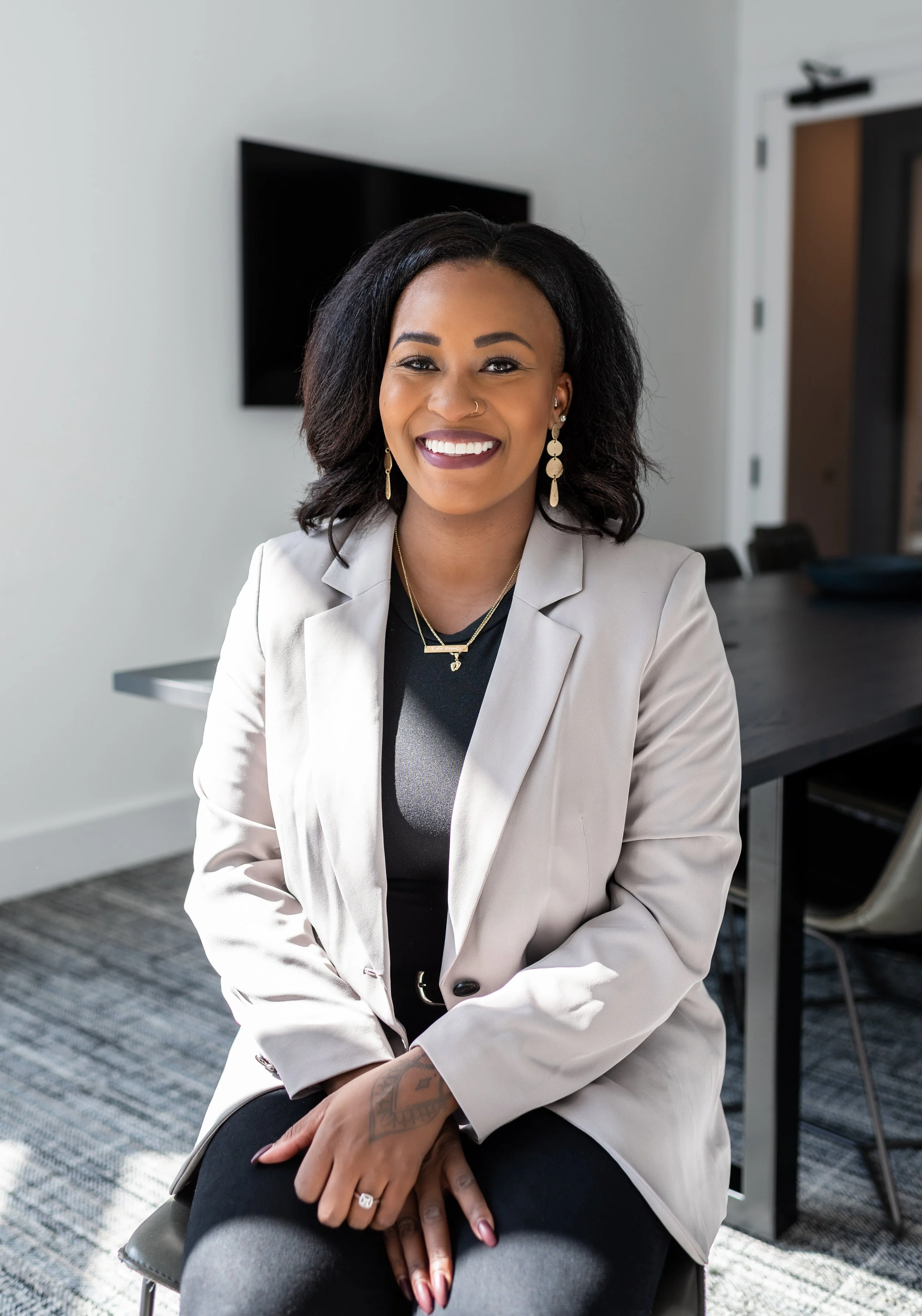 A woman with dark curly hair and a nose ring smiling, sitting in a conference room with a dark table and chair, wearing a light-colored blazer and jewelry.