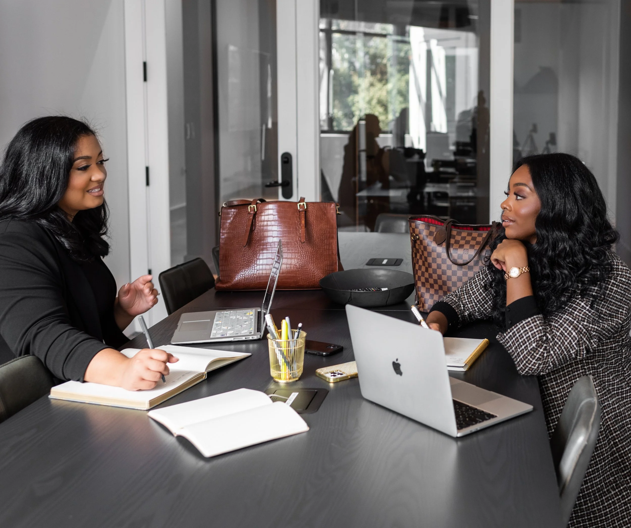 Two women having a business meeting in a conference room, sitting at a black table with laptops, notebooks, and bags.