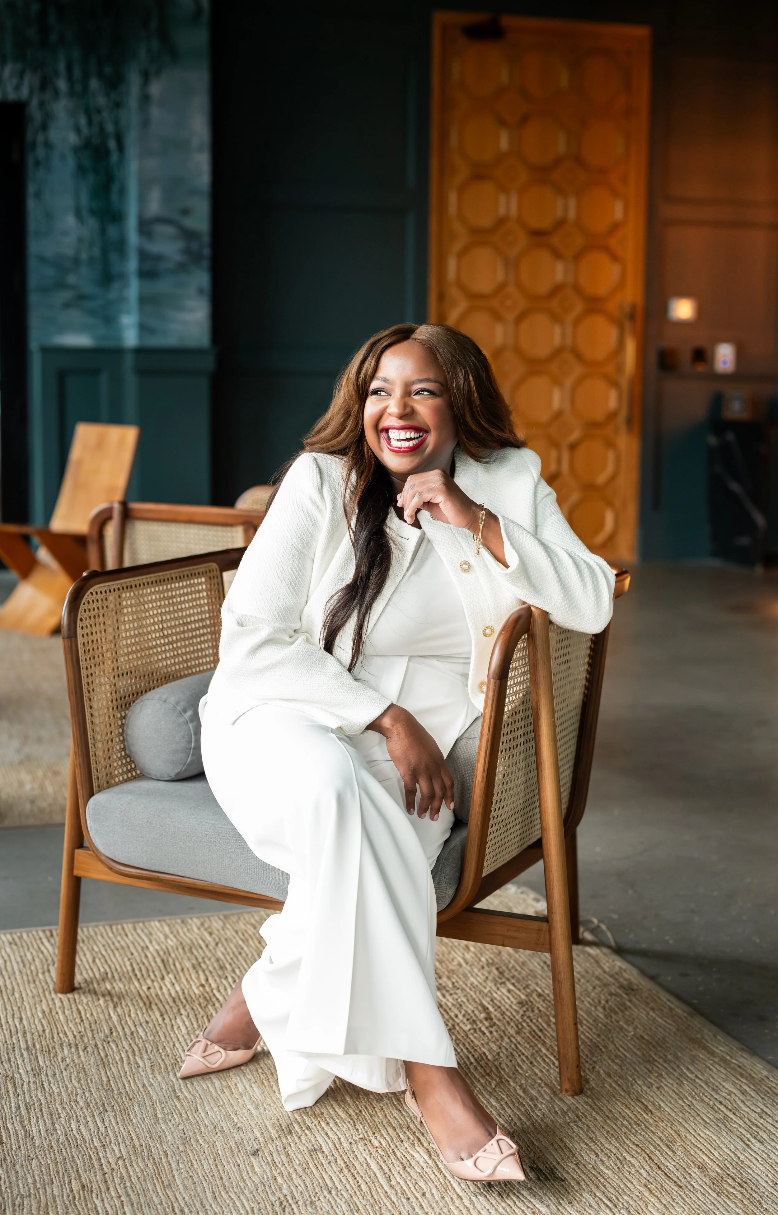 A woman in a white suit sitting on a modern wooden chair, laughing and smiling brightly in a stylish, contemporary room with wooden and dark green elements.