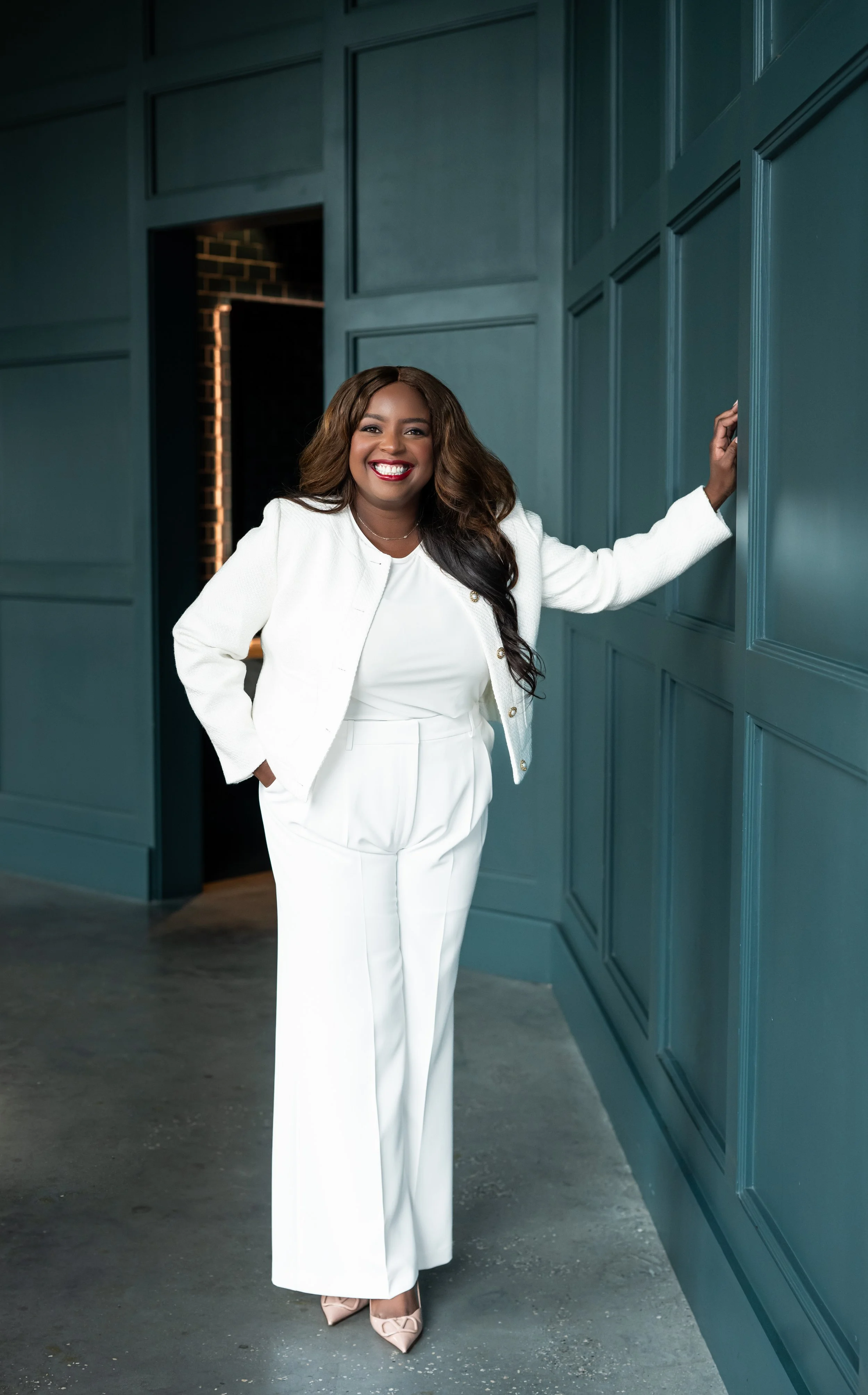 A smiling woman in a white suit standing against a teal paneled wall, with her right hand on her hip and her left hand touching the wall.