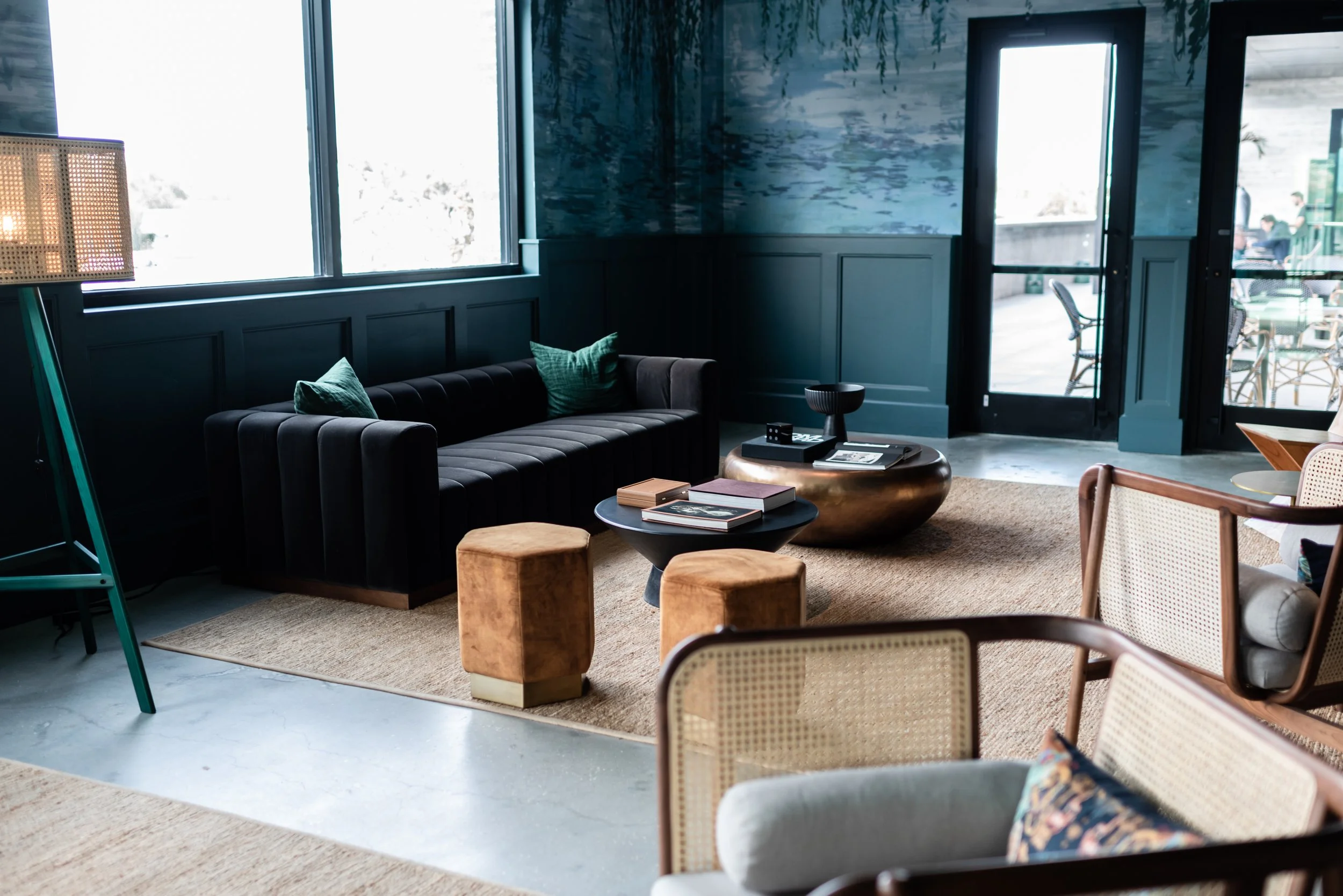 Cozy living room with black tufted sofa, green pillows, round coffee table, two brown stools, and an open door leading outside to a patio with outdoor furniture.