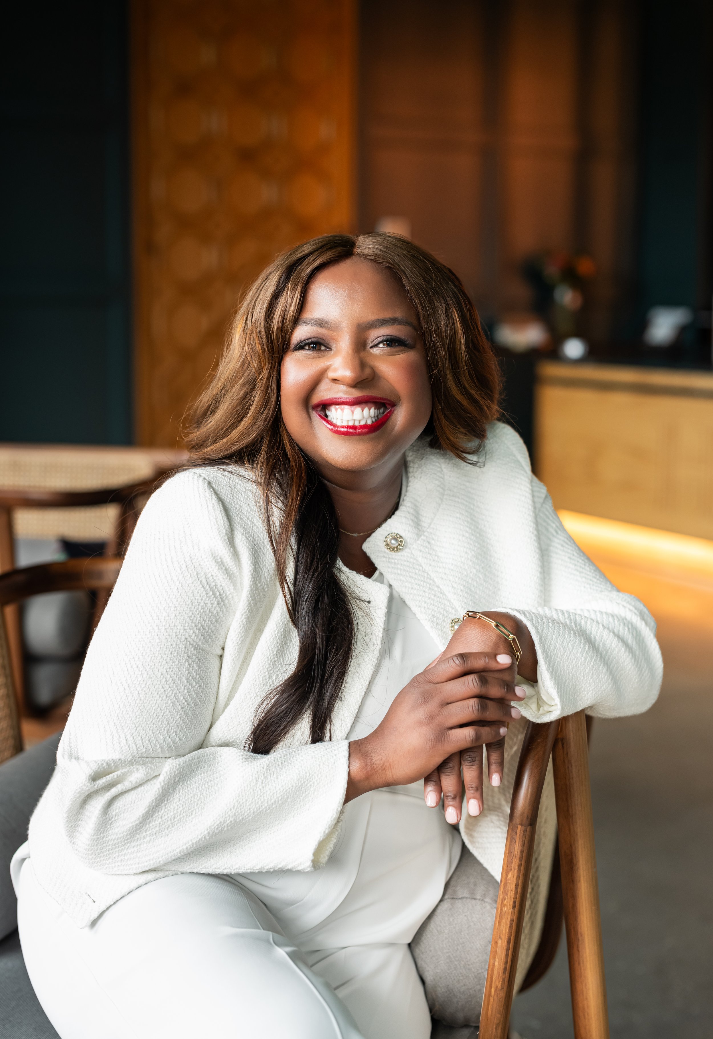 A smiling woman with dark brown hair in loose waves, wearing a white blazer, sitting on a chair, leaning on the backrest with one hand, in a warmly lit indoor setting.