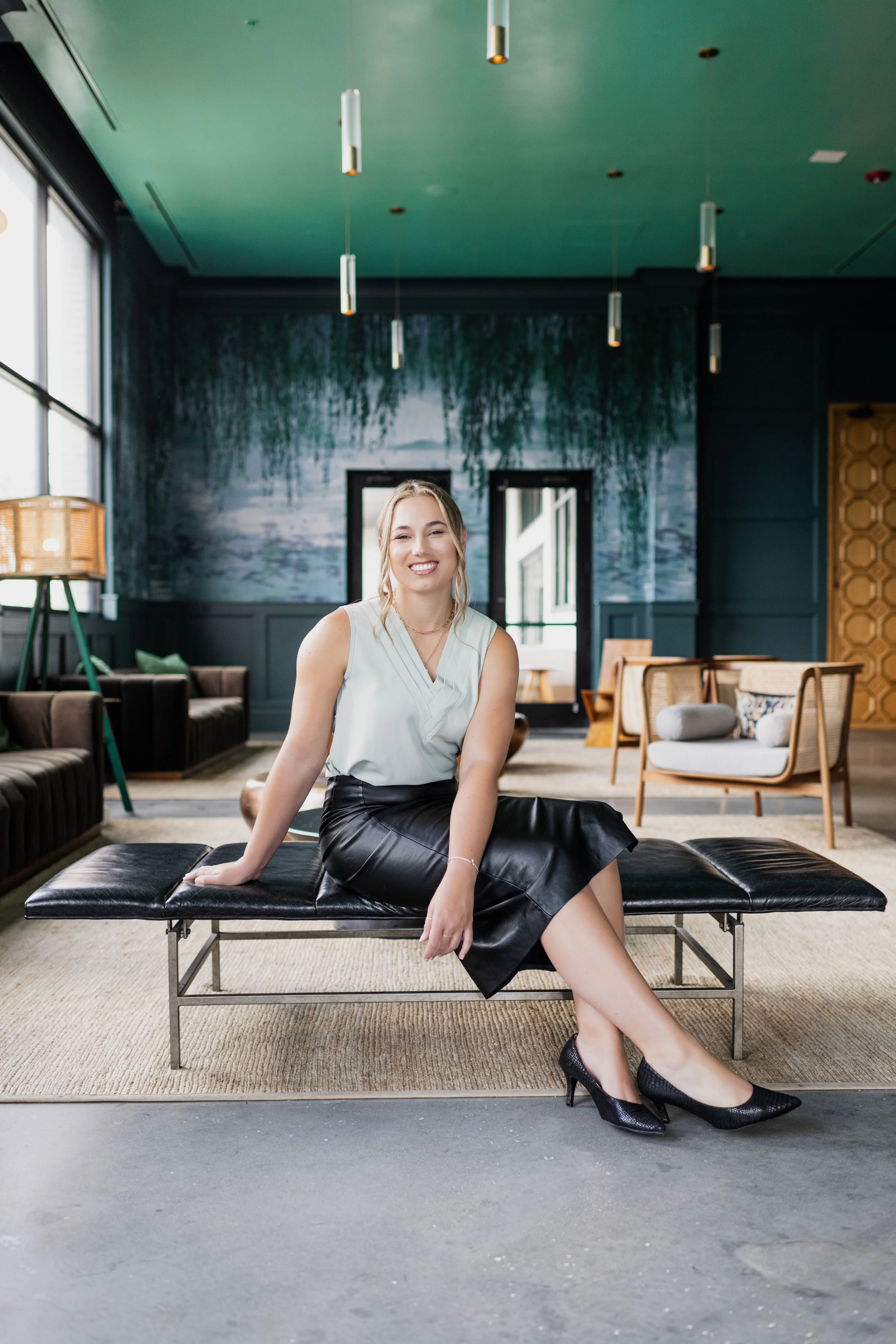 A woman sitting on a black leather bench in a stylish, modern lounge area with green walls, large windows, and contemporary furniture, smiling at the camera.