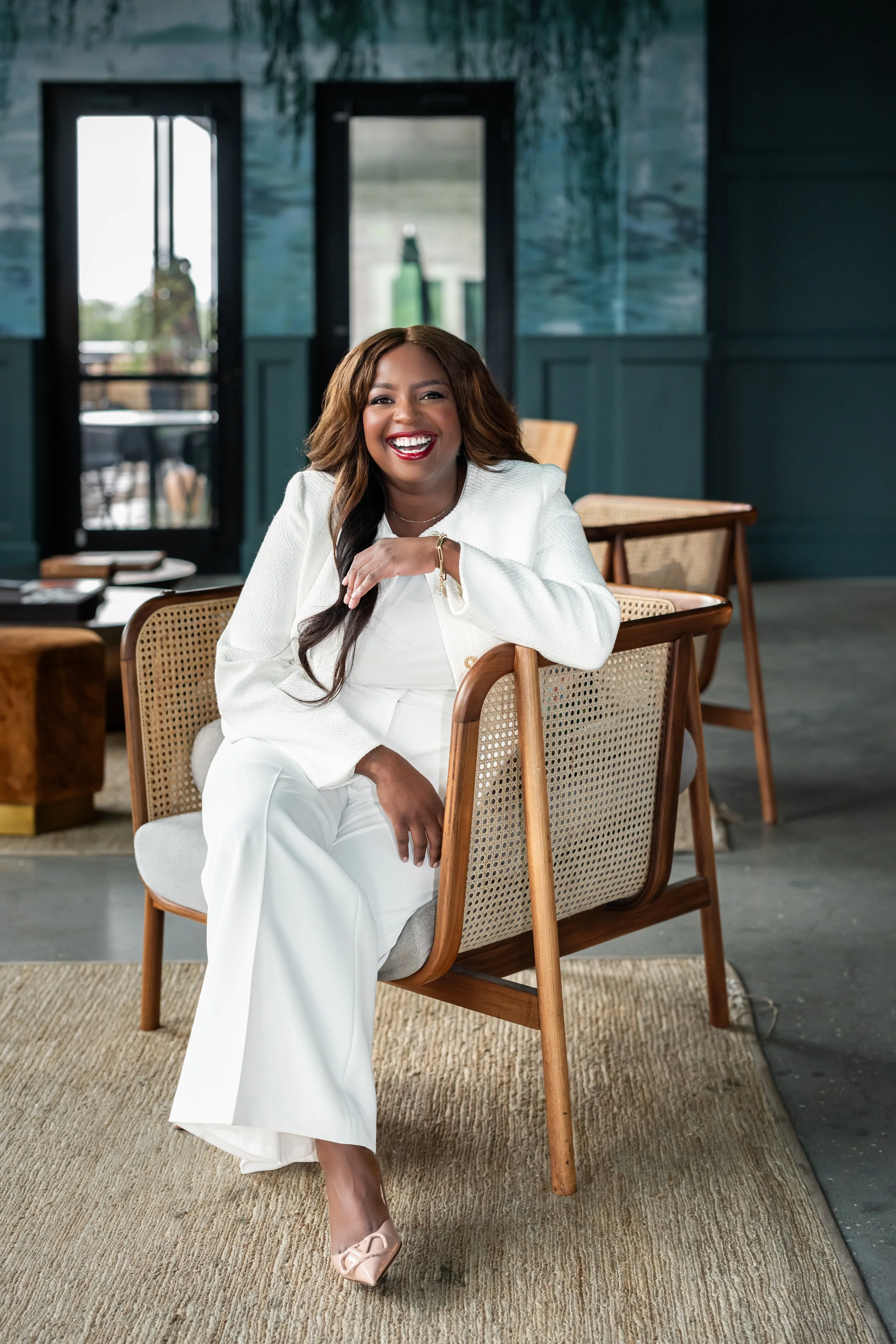 A woman with long brown hair wearing a white outfit, sitting on a wooden chair with woven backrest, smiling and laughing indoors.