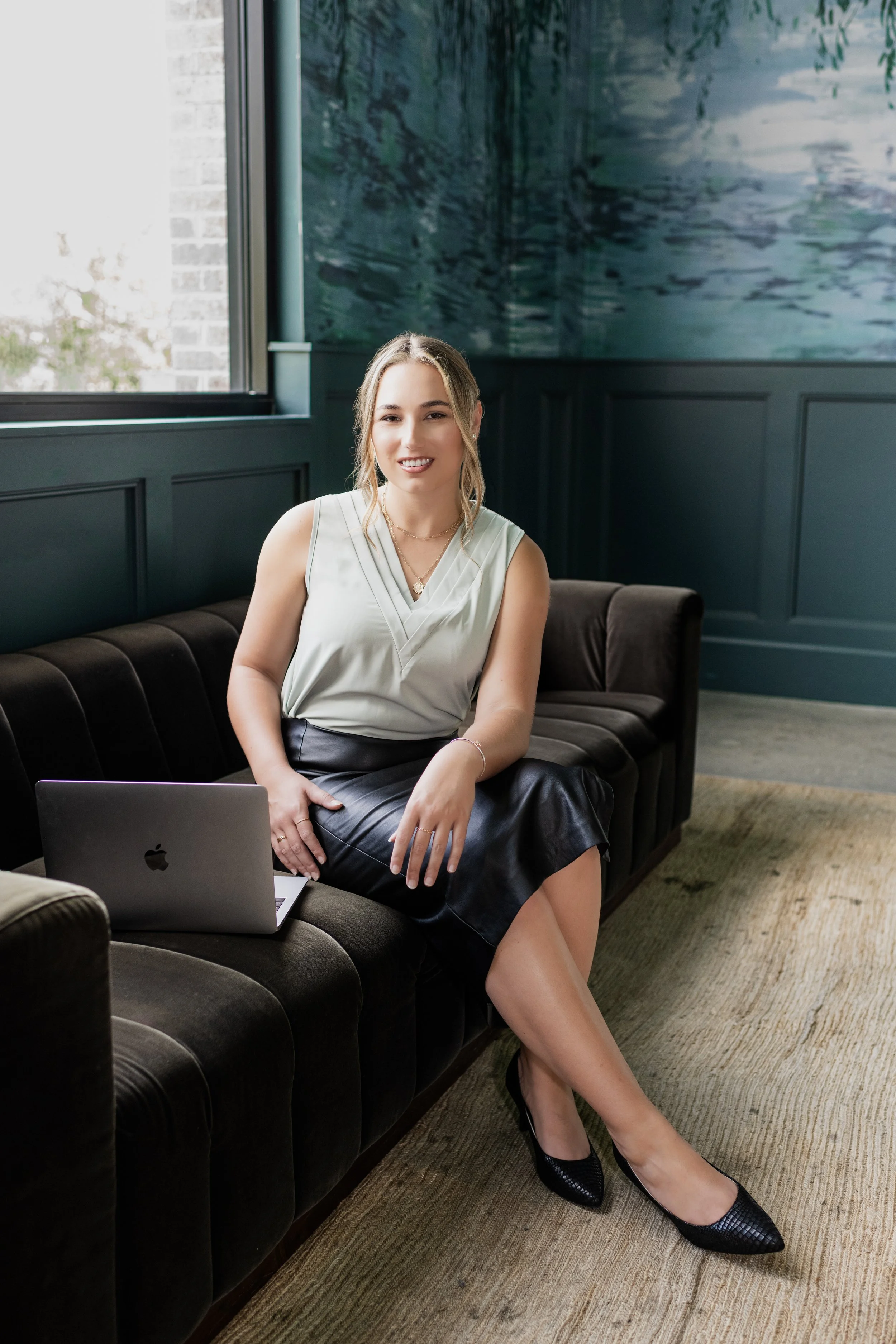 A woman sitting on a dark brown velvet couch with a silver laptop on her lap, in a stylish room with blue walls and a large window.
