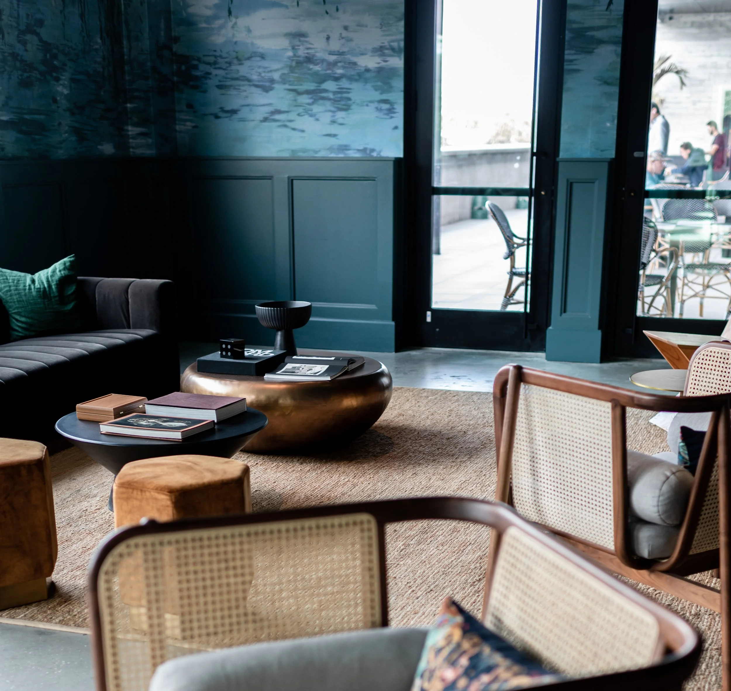Interior view of a stylish living room with a dark sofa, round coffee tables, and a patterned rug. Large glass doors lead to an outdoor seating area with tables and chairs.