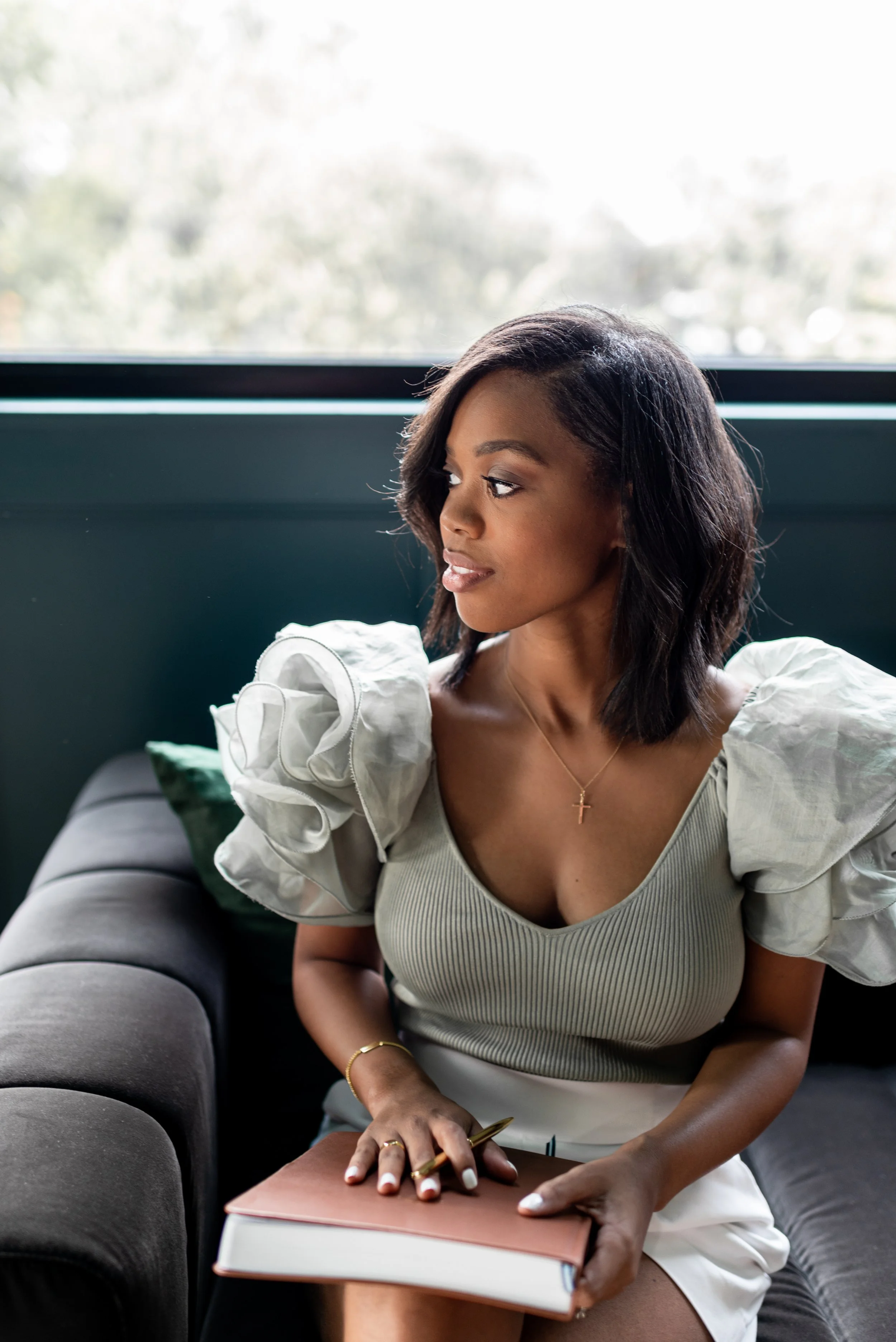 A woman with shoulder-length dark hair wearing a beige top with puffed sleeves, sitting on a dark sofa near a window, holding a notebook and pen, looking to the side.