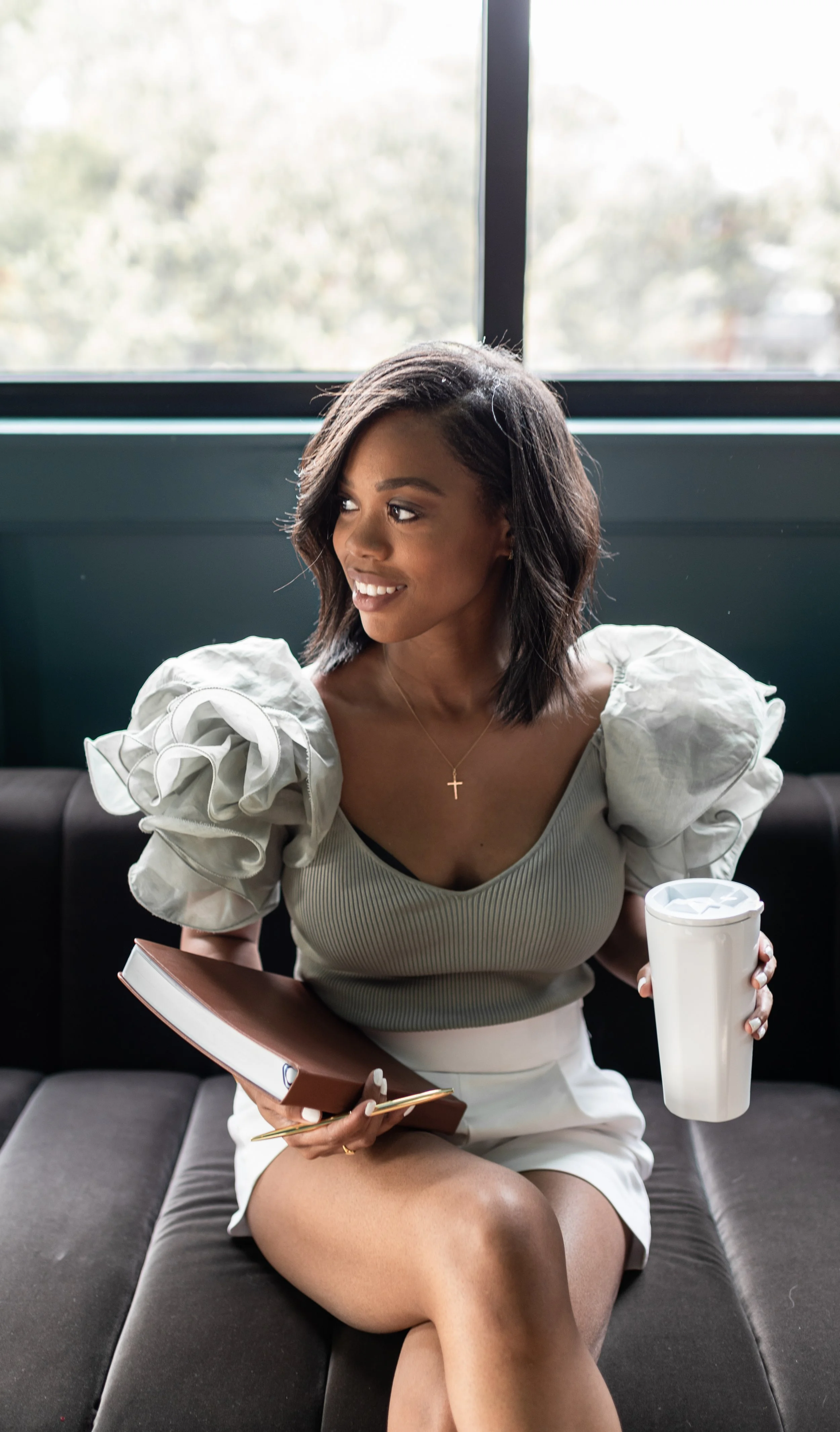 A young woman with shoulder-length black hair, wearing a gray top with puffed sleeves and a white skirt, sits on a black sofa holding a notebook and a pen in one hand and a white tumbler in the other. She is smiling and looking to her left, with a la