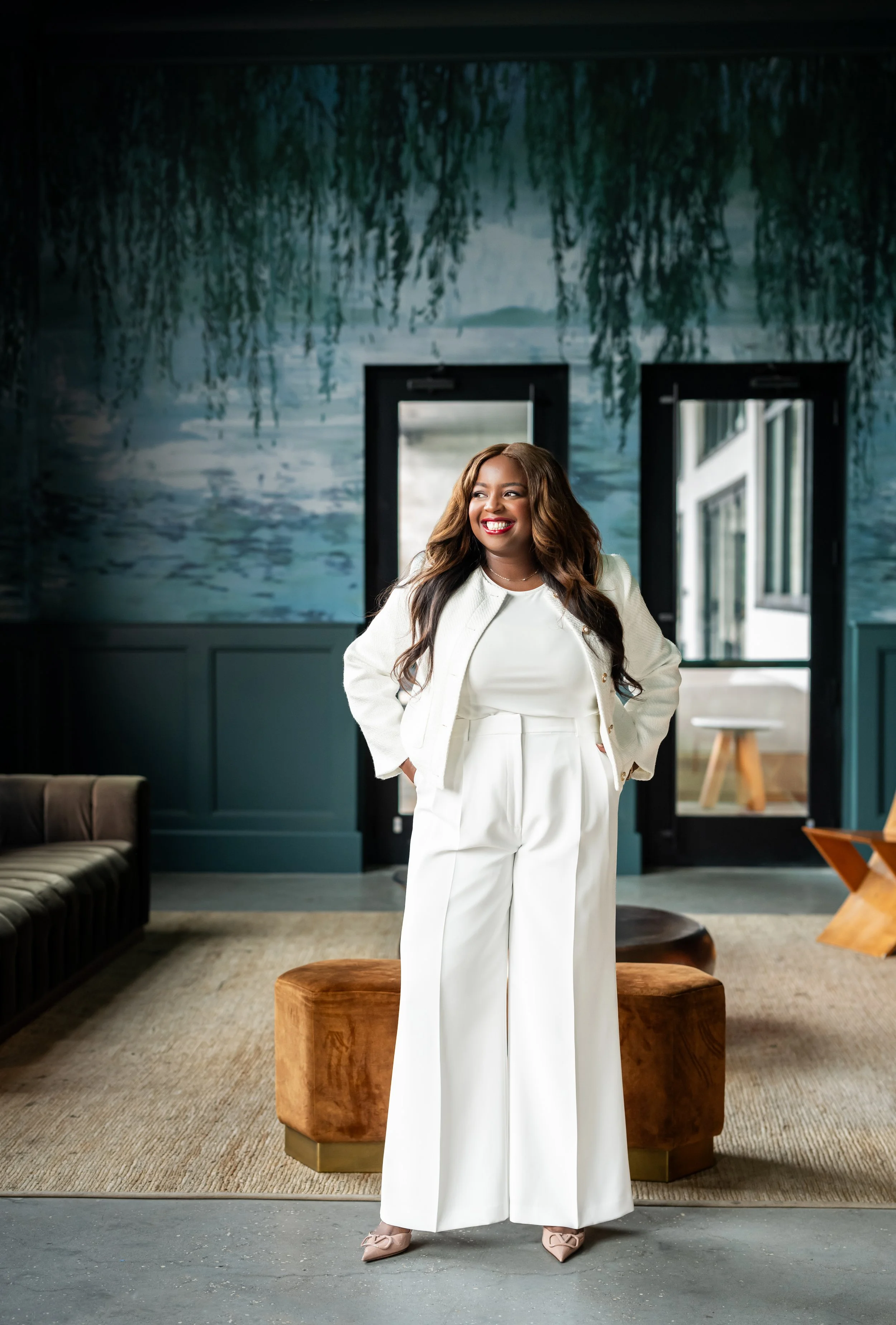 A woman with long, wavy hair and a bright smile, dressed in an all-white outfit, stands in a stylish indoor space with modern decor, including a mural and large windows.