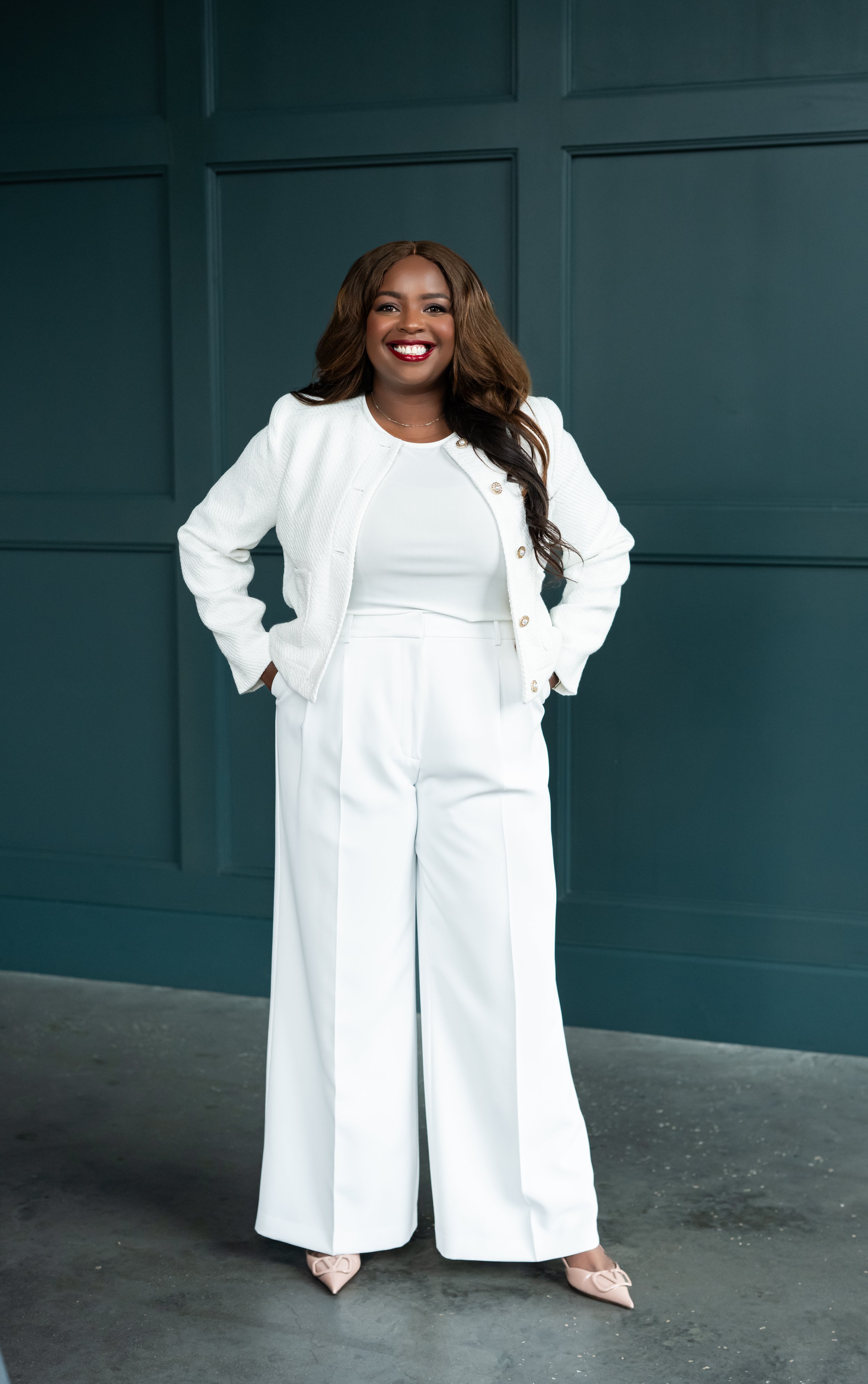 A woman with long brown hair, wearing a white suit and heels, standing with hands in her pockets and smiling in front of a dark blue paneled wall.
