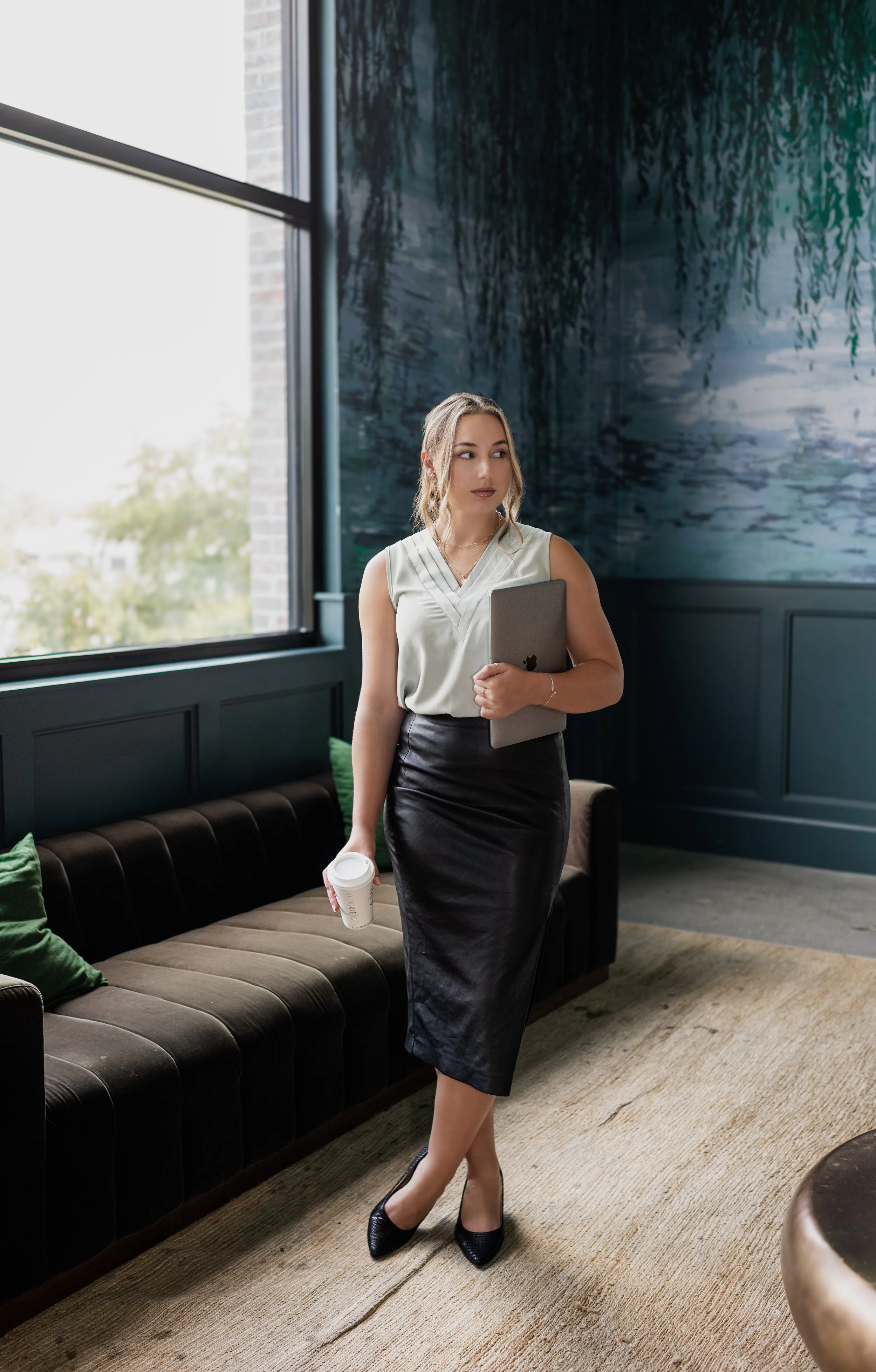 A woman in business attire holding a laptop and a takeout coffee cup, walking inside a modern office lounge with large windows, a dark leather couch, green pillows, and a large abstract wall painting.
