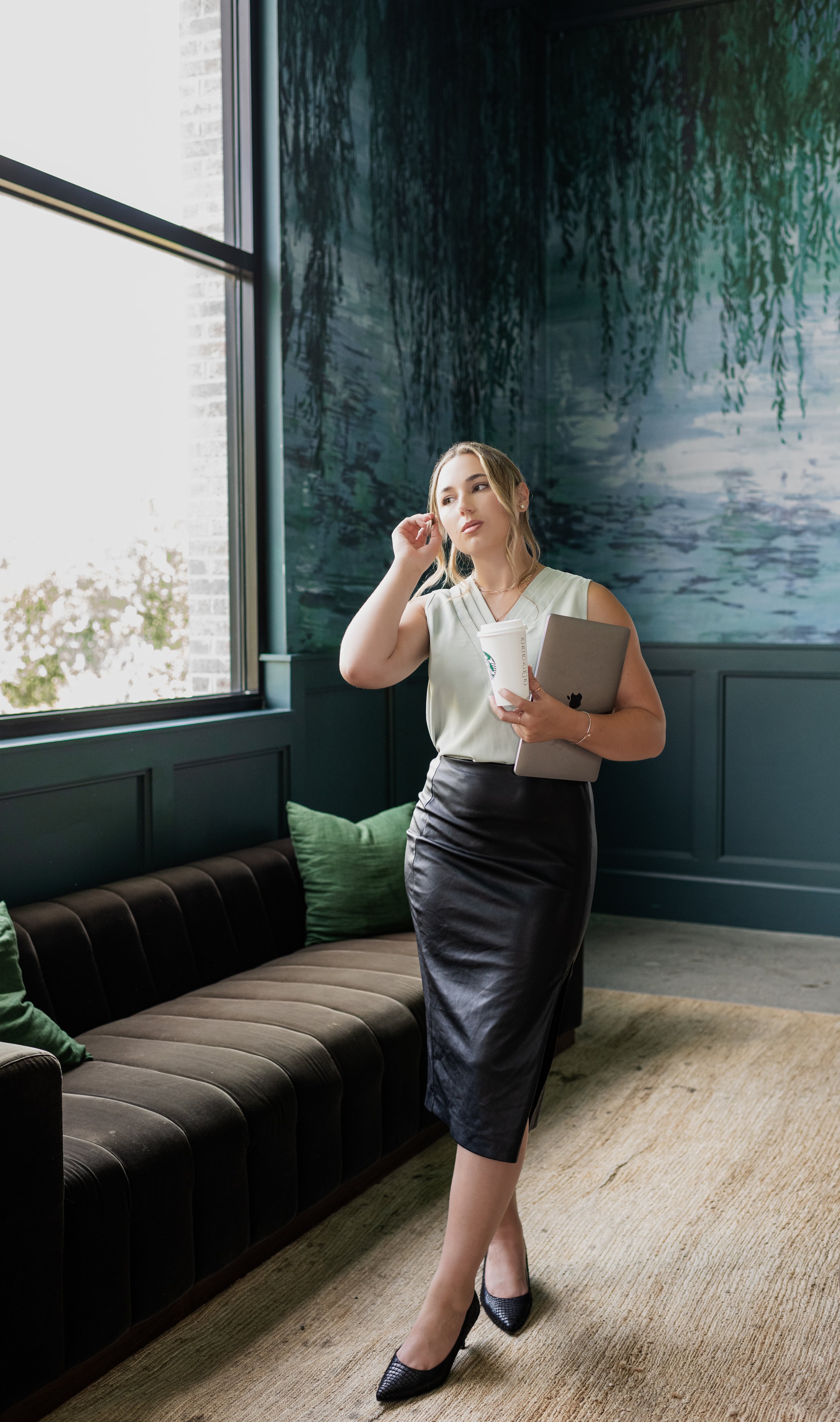 A woman in a cream blouse and black leather skirt stands by a large window in a modern cafe, holding a coffee cup and laptop, with green cushions on a black velvet sofa behind her.