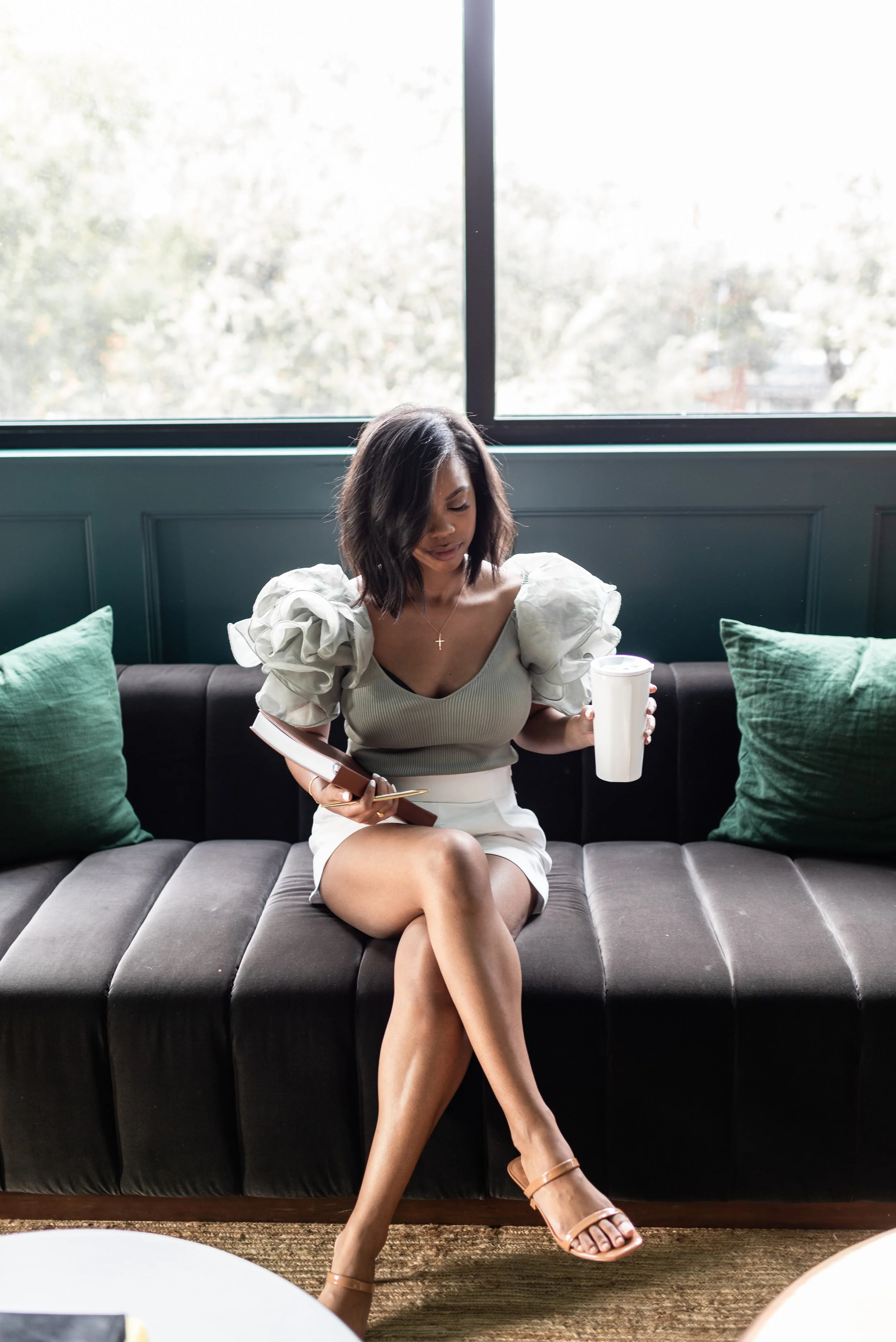 A woman sitting on a black sofa with green pillows, wearing a white blouse with puffed sleeves, holding a cup and a notebook with a pen, in front of a large window.